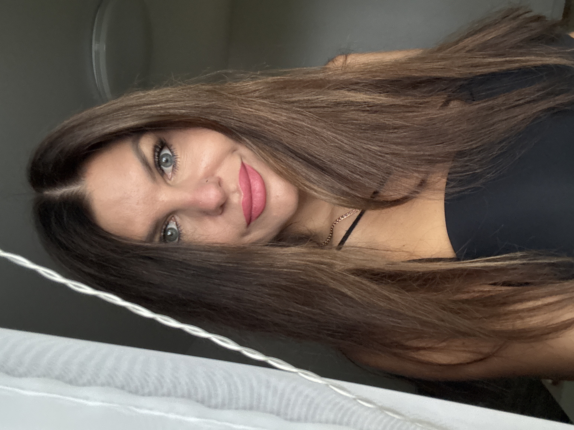 A woman with long, brown hair and blue eyes is looking at the camera. She is wearing a black top and a gold chain necklace. She has on pink lipstick and a small piercing on her nose. She is standing in front of a white curtain and a dark gray wall. There is a light fixture visible on the ceiling in the background. The image appears to be a selfie taken indoors.