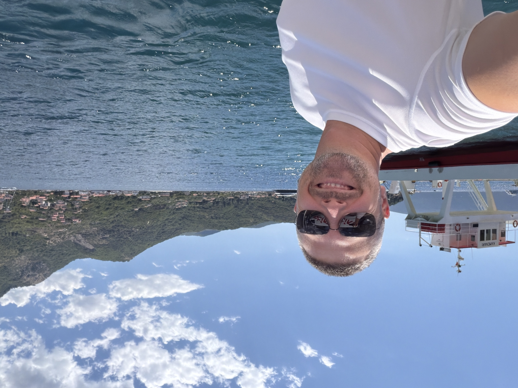 A selfie captures a smiling, fair-skinned man with short gray hair and a goatee, wearing a white t-shirt and dark sunglasses. He is on a boat in the water, with a ferry named "KAMENARI" visible to his left. The background shows a picturesque landscape of a bay with a town nestled on a green, mountainous hillside under a bright blue sky with scattered white clouds. The water is a deep blue, reflecting sunlight.