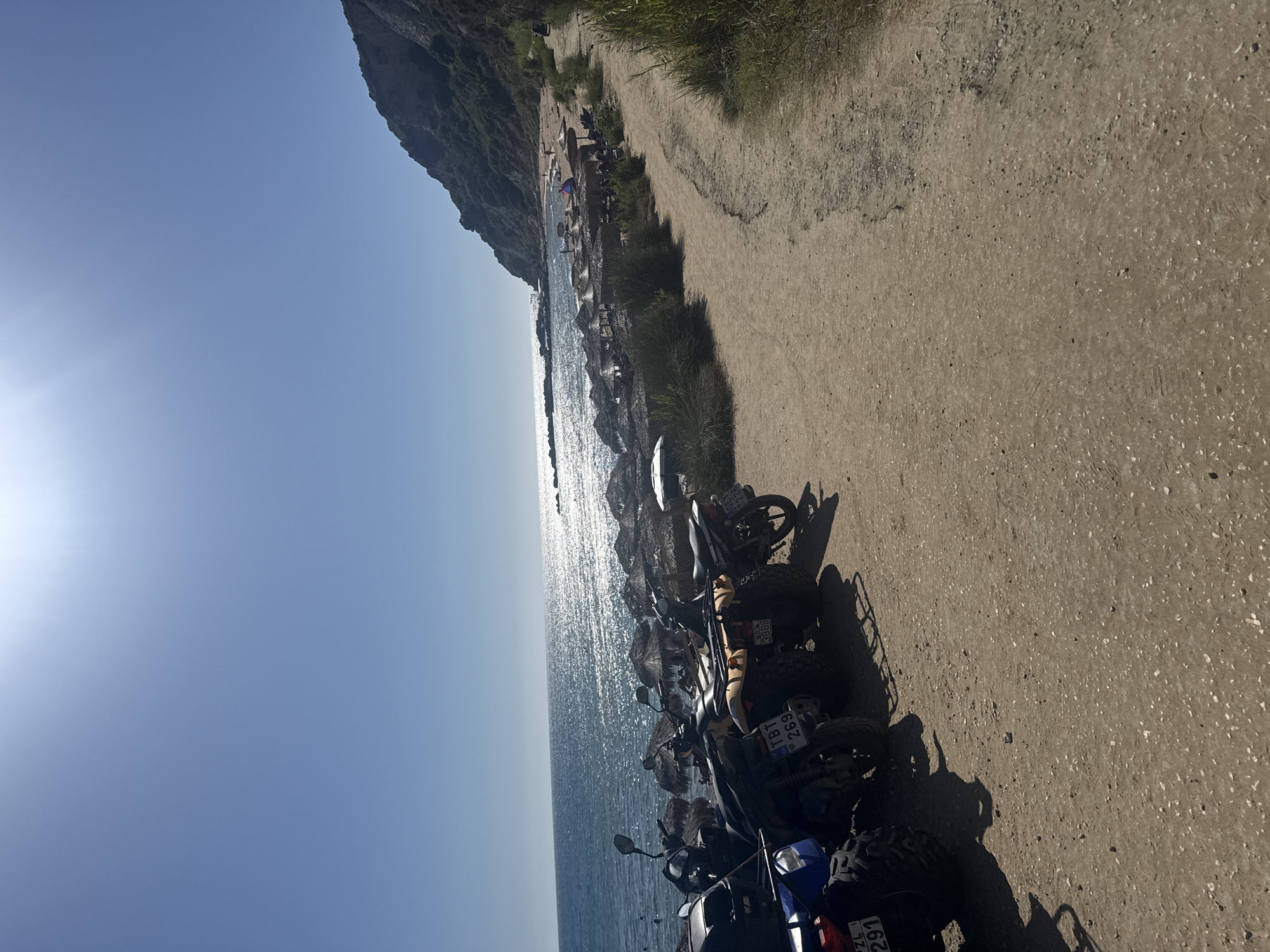 A sunny day at a beach in the Municipal Unit of Lefkimmi, Greece, features a row of parked ATVs and scooters on a dirt path leading to the shoreline. The sea is a vibrant blue, reflecting the bright sunlight, with small waves lapping at the shore. A stretch of beach is visible, dotted with straw umbrellas and a few sunbathers. In the background, a lush green hillside rises steeply, meeting a clear blue sky. The path is sandy and uneven, with vegetation growing along its edges.