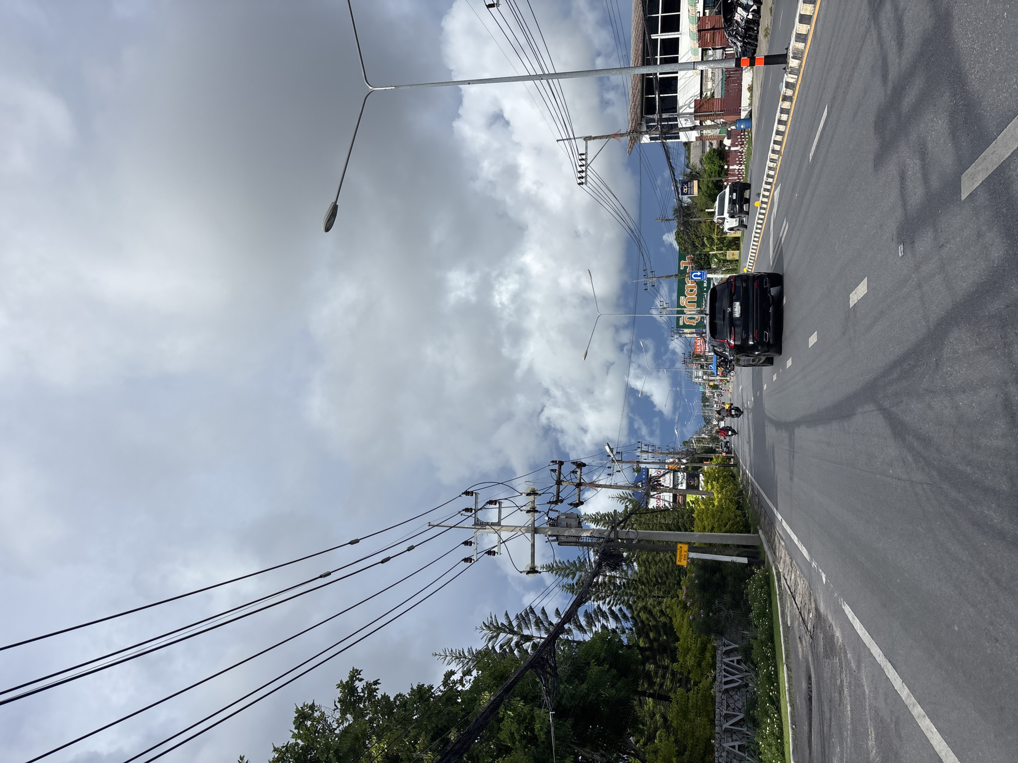 This is a photograph taken on a street in Rawai, Thailand, under a cloudy sky. The perspective is from a vehicle moving down the road. On the left side of the road, there is a wall with the inscription "A NEW WAY" and landscaping with white flowers and green foliage. Behind this, there are pine trees and other greenery. Utility poles with numerous wires stretch across the upper portion of the image, and a street lamp is visible on the right.
The road itself is asphalt with white lane markings. Several vehicles are present: a black SUV is in the foreground, followed by a white pickup truck. Further down the road, two motorcycles are visible. The buildings on the right appear to be commercial establishments, with one featuring a white facade and dark windows. The overall scene depicts a typical street view in a coastal town.