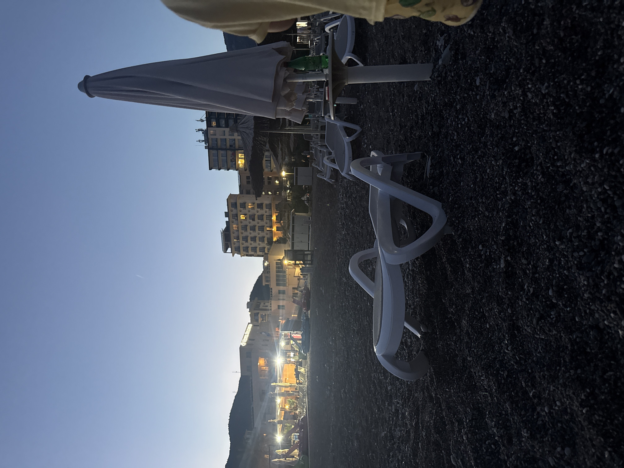 The image depicts a beach in Boreti, Montenegro, at dusk. In the foreground, a white plastic lounge chair is positioned on a dark, pebbly beach. Behind it, several other lounge chairs and closed beach umbrellas are visible, some arranged around tables. A white, closed umbrella stands prominently to the right, with a green bottle placed on a nearby table. In the background, buildings with illuminated windows suggest a resort or hotel area, with mountains forming the backdrop. The sky is a clear, deep blue, transitioning from lighter hues near the horizon. A thin contrail is visible in the upper portion of the sky. The overall atmosphere is serene and quiet, indicative of the end of the day.