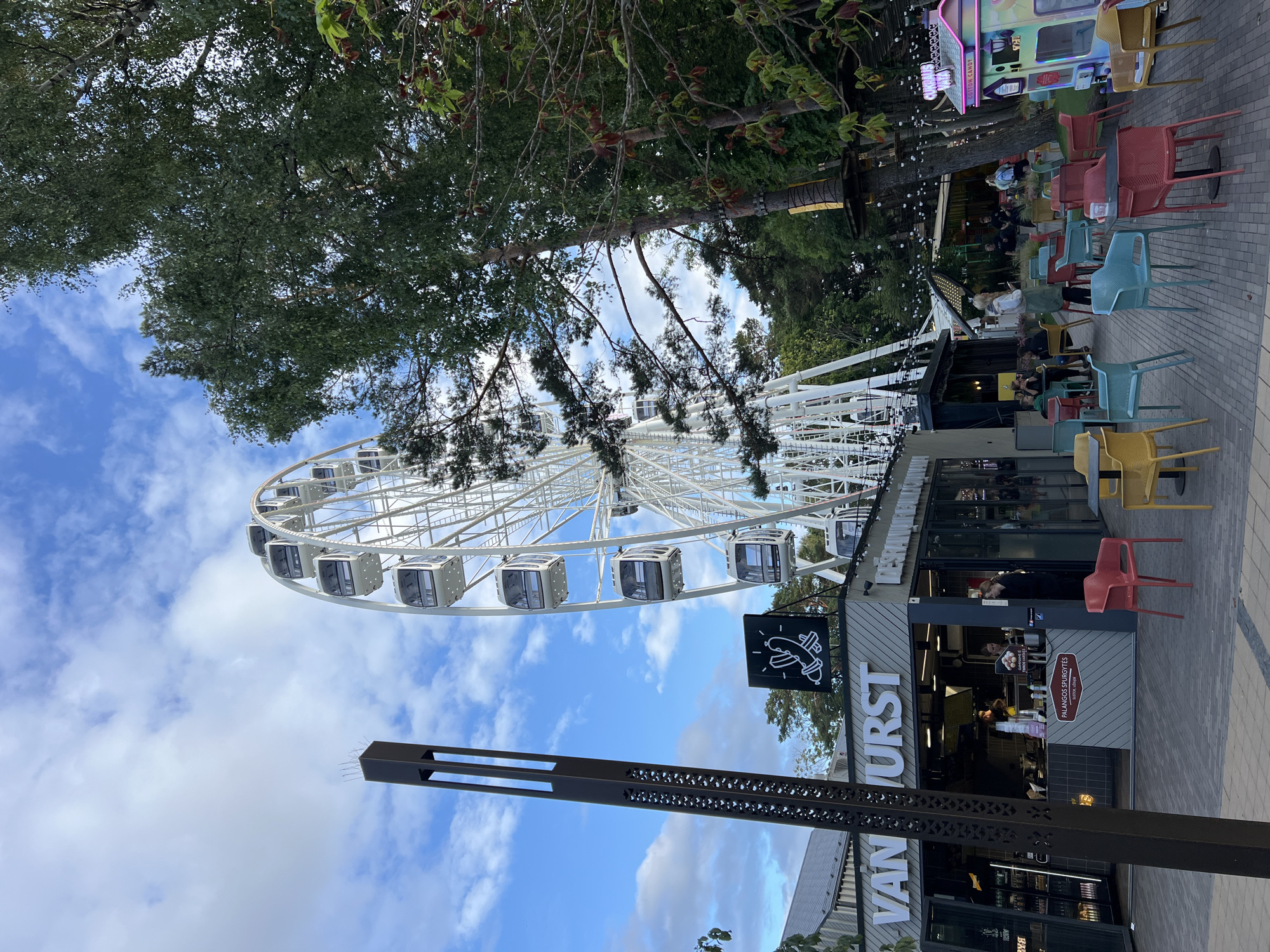 The image depicts an outdoor scene in Palanga, Lithuania, likely at an amusement or entertainment area. A large white Ferris wheel dominates the background, with its gondolas visible against a cloudy blue sky. In the foreground, a food stall with the signage "VAN URST" is prominent. People are visible inside the stall and standing at a counter. Outside the stall, an array of colorful chairs and tables are set up on a paved area. To the left of the food stall, a tall, dark, decorative pole stands. Further in the background, trees and what appears to be a ropes course are visible, along with strings of lights. A colorful cotton candy machine is also present on the right side of the frame. The overall atmosphere suggests a lively outdoor leisure venue.