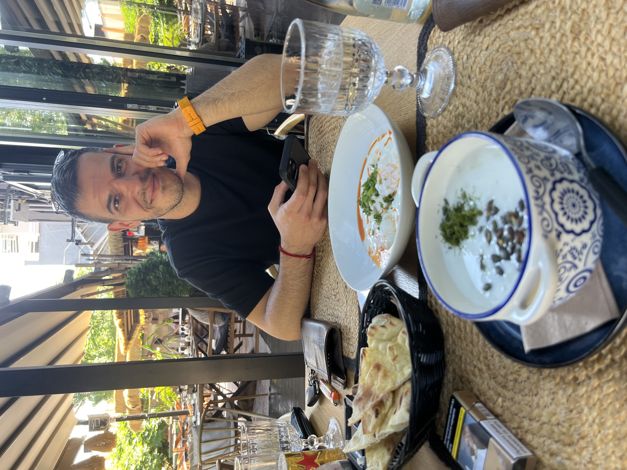 A man sits at an outdoor restaurant table with food and drinks. He is looking at his phone. The table is set with a bowl of soup, a plate of flatbread, glasses of water and beer, a wallet, keys, and a pack of cigarettes. The man is wearing a black t-shirt and an orange watch. The restaurant appears to be located in Sofia, Bulgaria, with outdoor seating and lush greenery visible in the background.