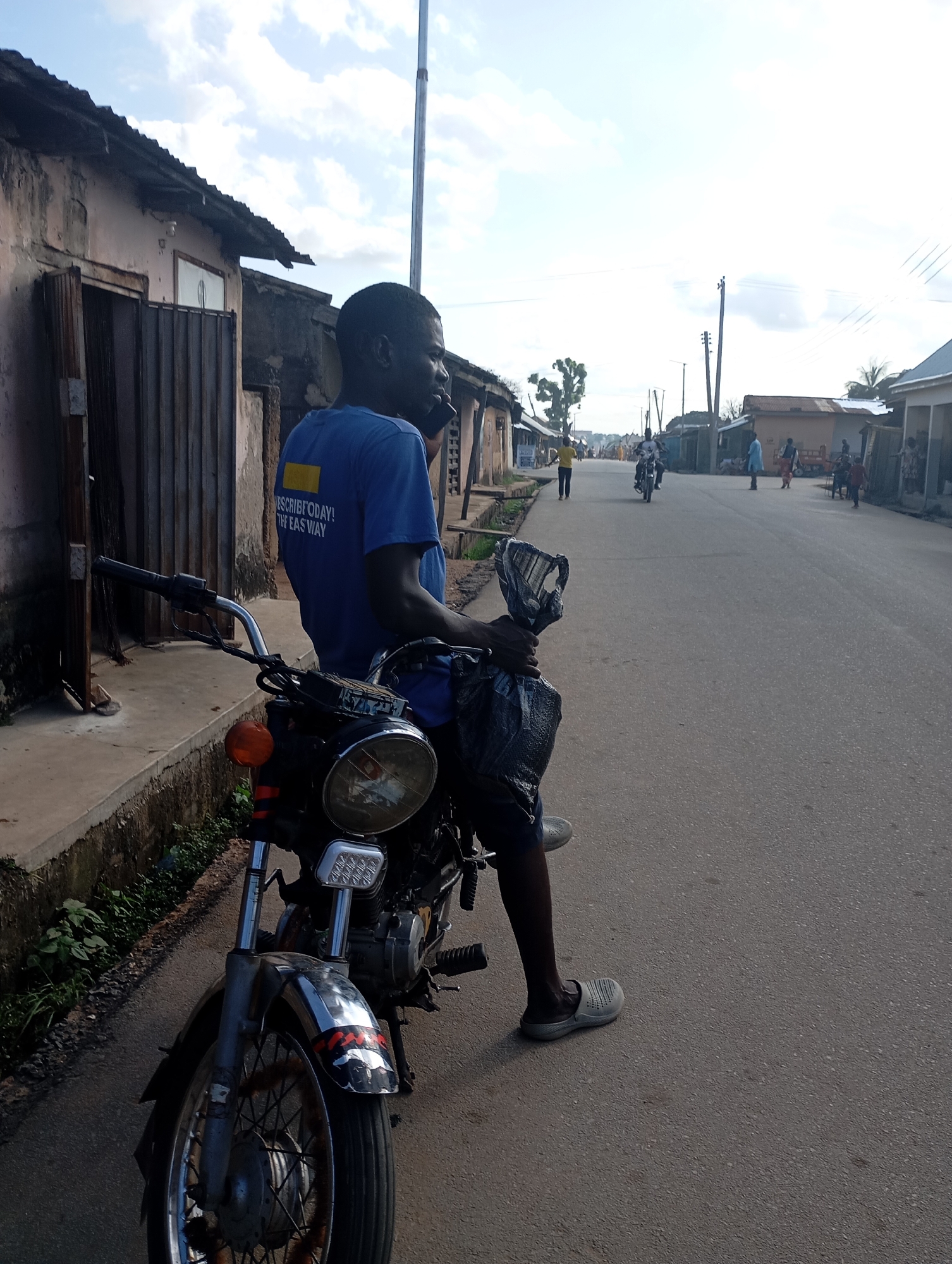The image depicts a street scene in Shendam, Nigeria. In the foreground, a young man wearing a blue t-shirt with yellow text on the back, "SUBSCRIBE TODAY! THE EASY WAY", is seated on a motorcycle. He is holding a black plastic bag in his right hand. The motorcycle is dark-colored with chrome accents, including the handlebars, headlight, and wheel spokes. The street is paved and appears to be a road in a town or city. Buildings line both sides of the street, with some appearing to be shops or residences with corrugated metal roofs. In the background, other people and a few more motorcycles are visible, suggesting a moderately active street. The sky is partly cloudy. The lighting indicates it is daytime, likely in the morning or late afternoon due to the angle of the sun casting shadows.