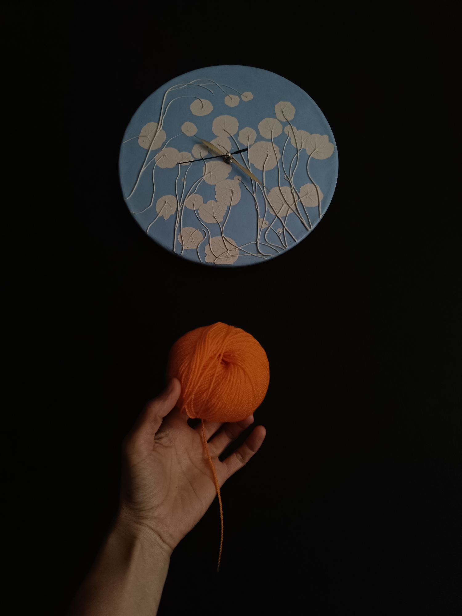 The image displays a minimalist, high-contrast composition against a deep black background. In the upper center, a light blue circular wall clock is the primary subject. Its face is adorned with raised, light cream-colored botanical patterns, resembling stylized leaves or seed pods on flowing stems, giving it a textured, artistic appearance. Gold-toned hour and minute hands indicate an approximate time of 1:50 or 2:50.
Positioned centrally in the lower half of the frame, a human hand, likely an adult's, extends from the left. The hand, with visible skin texture, holds a vibrant, perfectly wound ball of bright orange yarn or thread. A single strand of yarn trails downwards from the ball.
The scene appears to be an indoor, staged setting, possibly a studio, with the dark background serving to emphasize the colorful objects. There is no specific activity beyond the hand holding the yarn, suggesting a still life or a display of these items. No visible text or explicit location cues are present within the image itself, though the context provided is Lermontov, Russia. The lighting is focused on the subjects, allowing the dark background to create a strong visual contrast.
