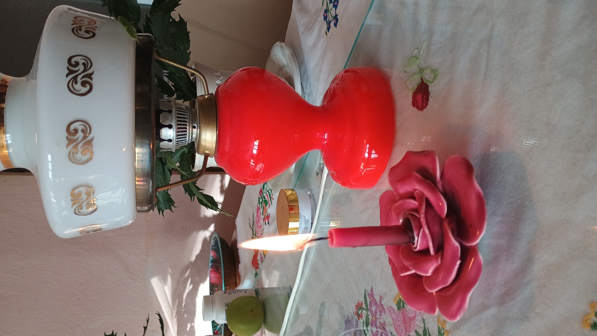 In Sokolov, Czechia, a close-up shot captures a still life arrangement on a tablecloth. The foreground features a lit pink candle in a rose-shaped holder, its flame casting a warm glow. To the right, a bright red oil lamp with a white glass shade, adorned with gold scrollwork, stands prominently. Behind the lamp, a potted plant is visible. On a glass surface between the candle and the lamp, a small white jar with a gold lid and a light green fruit are placed. The background is a soft pink wall.
