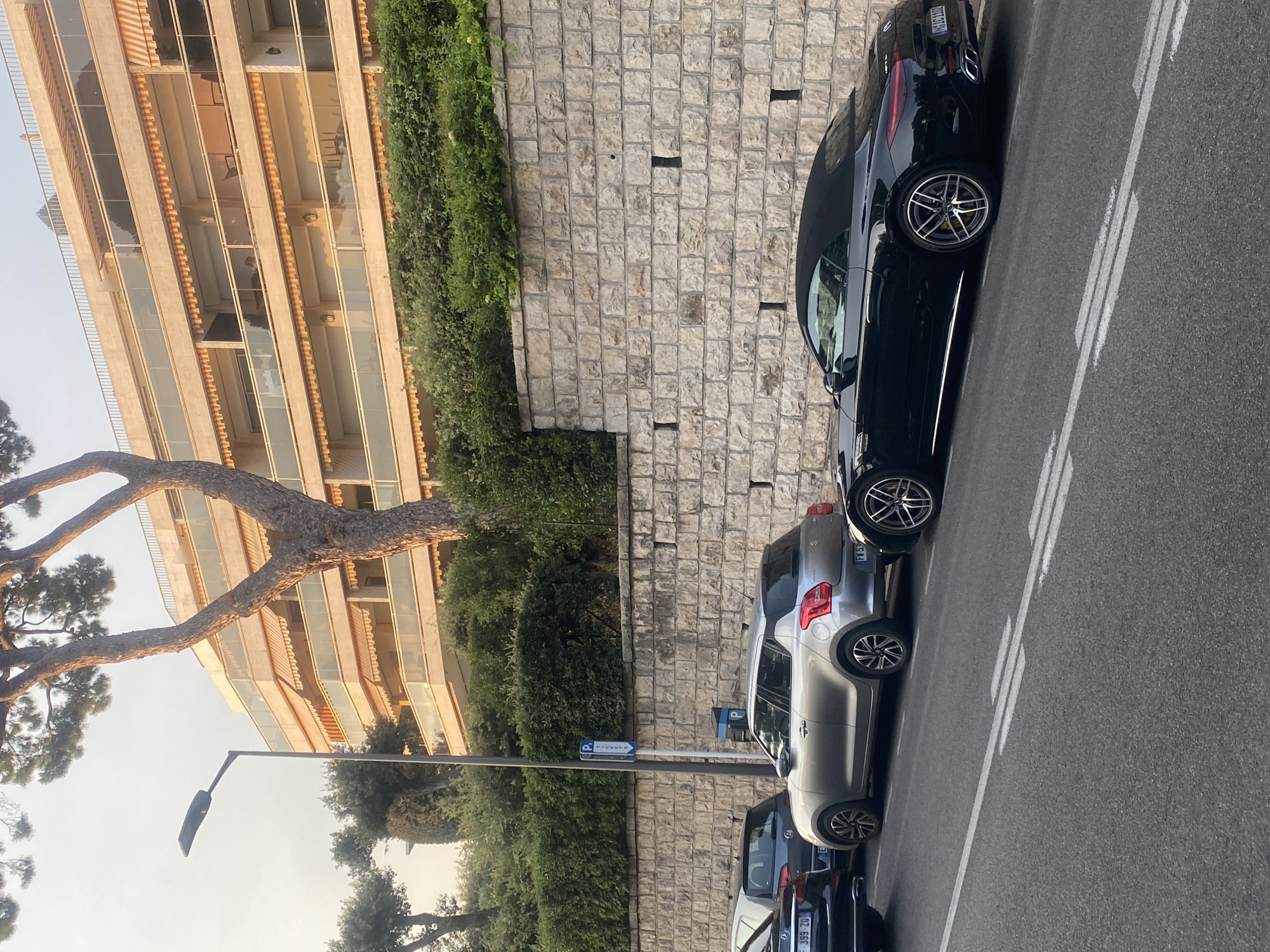 In the city of Antibes, France, on a street lined with parked cars, a multi-story building with balconies and awnings stands behind a stone wall topped with greenery. A street lamp is visible on the left, and a parking sign is mounted on a pole. The cars parked on the street include a black convertible, a silver hatchback, and other vehicles partially visible. The street surface is asphalt with white lane markings.