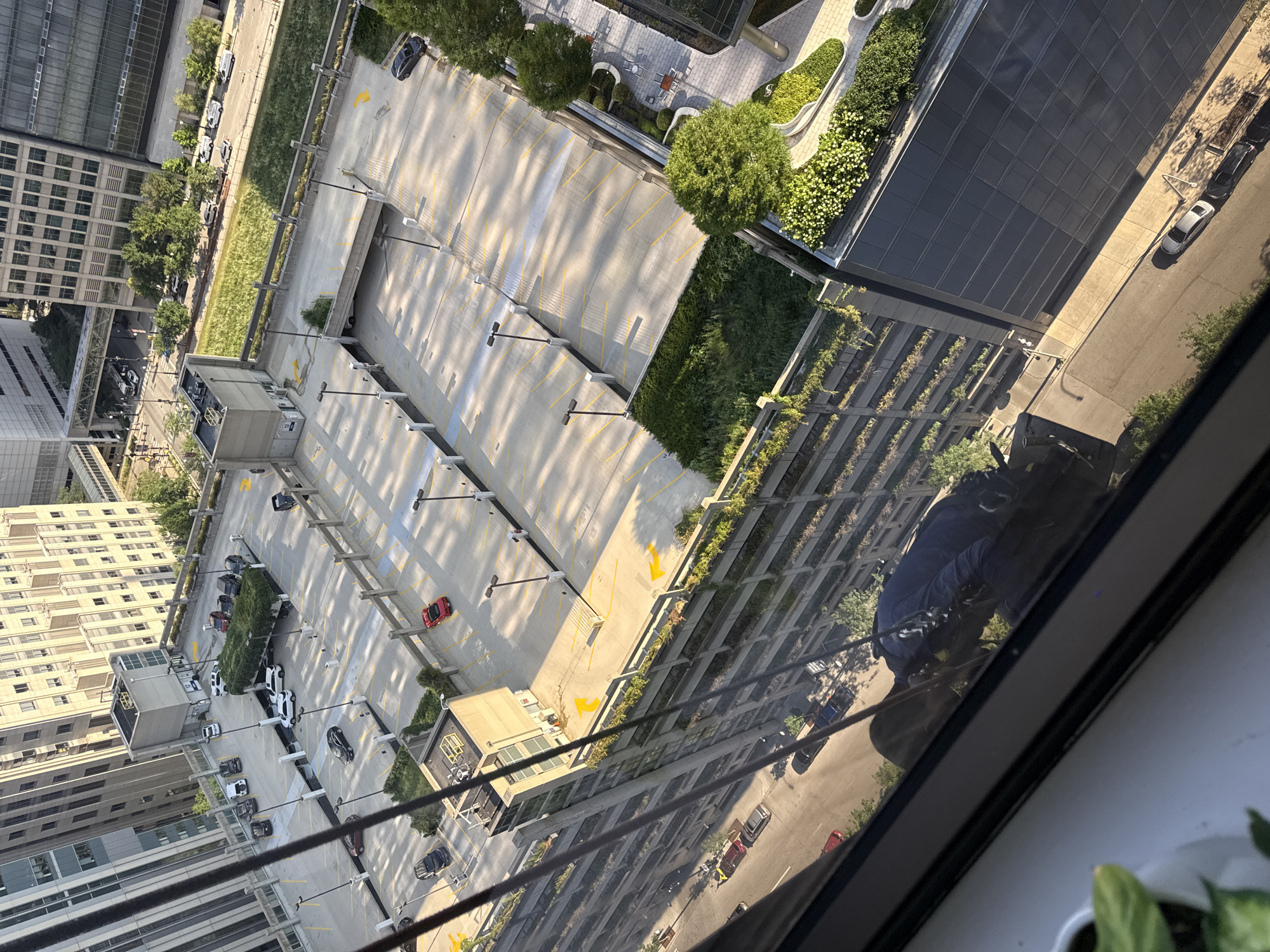 This is an aerial, high-angle photograph taken from a window overlooking a multi-level parking garage in Chicago, Illinois. The image captures various sections of the parking structure, including empty parking spots marked with yellow lines, concrete barriers, light poles, and decorative green landscaping on some levels. Several cars are visible parked on the different tiers of the garage, including sedans and SUVs in various colors such as black, white, silver, and red.
In the foreground, the reflection of a window and some indoor elements, including a potted plant and the reflection of a person in a dark uniform, are visible. The person appears to be wearing a harness and is possibly a window cleaner. The environment outside the window is characterized by urban architecture, with tall buildings surrounding the parking garage and a street running alongside it, also with parked cars and trees. The sunlight casts shadows across the parking lot, indicating it is daytime.