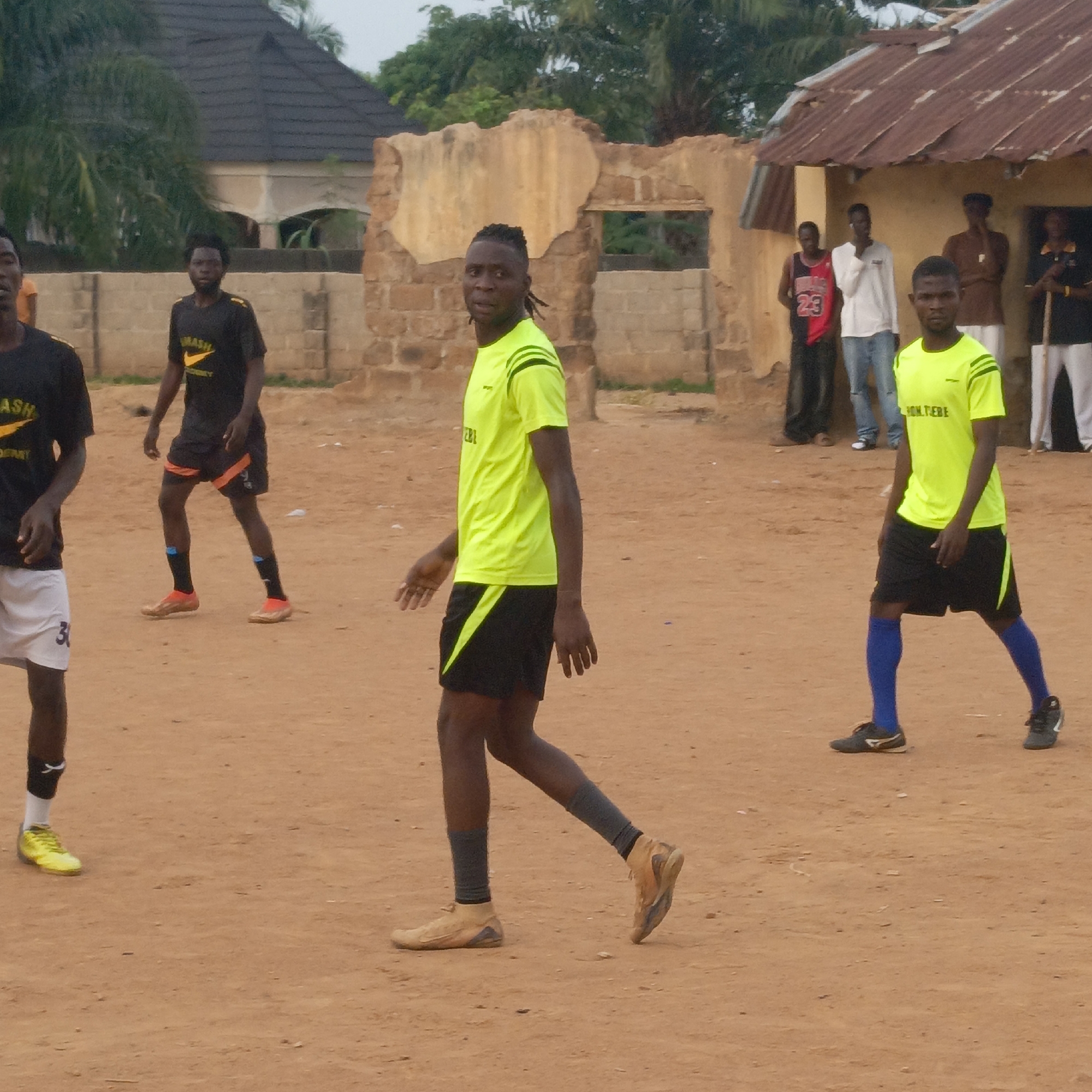 The image shows a group of men on a dirt field, possibly in Akwanga, Nigeria. Five men are dressed in athletic attire, suggesting they are playing or have just finished playing a sport. Two are wearing bright yellow and black uniforms with blue socks, while two others wear black uniforms with orange accents. One man wears white shorts and a black shirt. The athletes are in the foreground of the image, spread across the frame.
In the background, near what appears to be a weathered building, several more men are standing and watching. Some appear to be casually dressed. A damaged, partially ruined building with a missing roof is visible behind some of the men. Other buildings are in the far background. The ground is bare, reddish-brown dirt. The sky is overcast.