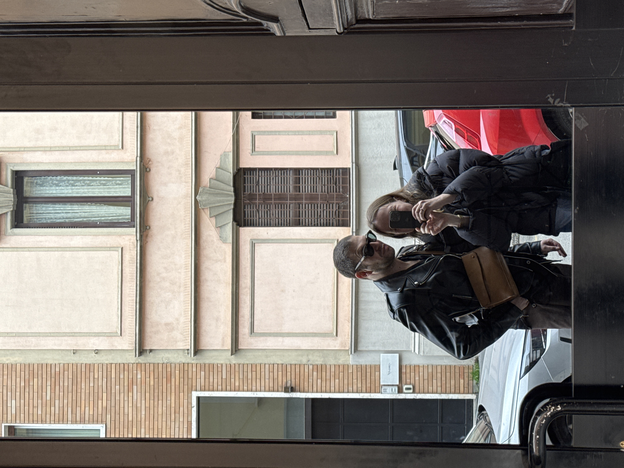 The image displays a reflection of two individuals and a city street scene, likely captured in a mirror or glass door in Milan, Italy.
A male subject, appearing to be in his 30s-40s, stands on the left. He has short dark hair and wears dark sunglasses, a black leather biker jacket over a dark shirt, brown trousers, and a light brown leather cross-body bag. His left hand is in his pocket, and he is positioned facing slightly to the right of the frame.
A female subject, appearing to be in her 20s-30s, stands on the right. She has long brown hair and wears a black puffer jacket over a striped garment. She holds a dark-colored smartphone horizontally at chest height, aimed towards the reflection, engaging in a selfie action. A gold-colored watch is visible on her left wrist.
The reflected background reveals an urban street. To the left, a building with light brown brickwork features a white-framed window and a dark grey ground-level door with a visible street number "0". Adjacent to this, a light pink stucco building exhibits classical architectural elements, including framed rectangular wall panels, cornices, and decorative fan-like motifs above windows. One window features dark frames and white lace curtains, while another is covered by dark wooden shutters (*persiane*). Two vehicles are visible parked along the reflected street: a silver sedan, partially obscured behind the male subject, and a red hatchback, partially obscured behind the female subject. The architectural style, particularly the light pink stucco and traditional *persiane*, is consistent with urban buildings found in Milan, Italy.