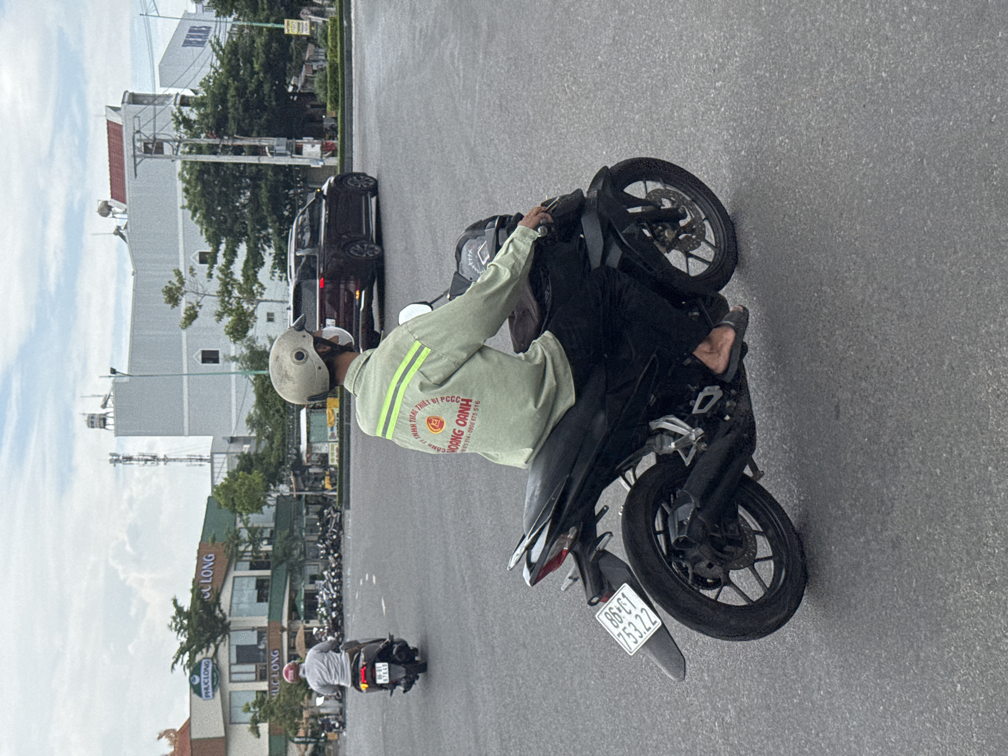 A man wearing a light green long-sleeved shirt with reflective stripes and black pants rides a black motorcycle on an asphalt road. He has a white helmet on and is wearing dark sandals. The motorcycle has a license plate that reads "86-C1 753.22". In the background, there are buildings, trees, and other motorcycles parked. One of the buildings has the text "PHUCLONG" and "CUULONG" on it. The sky is overcast. This scene appears to be in the city of Phan Thiết, Vietnam, as indicated by the license plate format.
