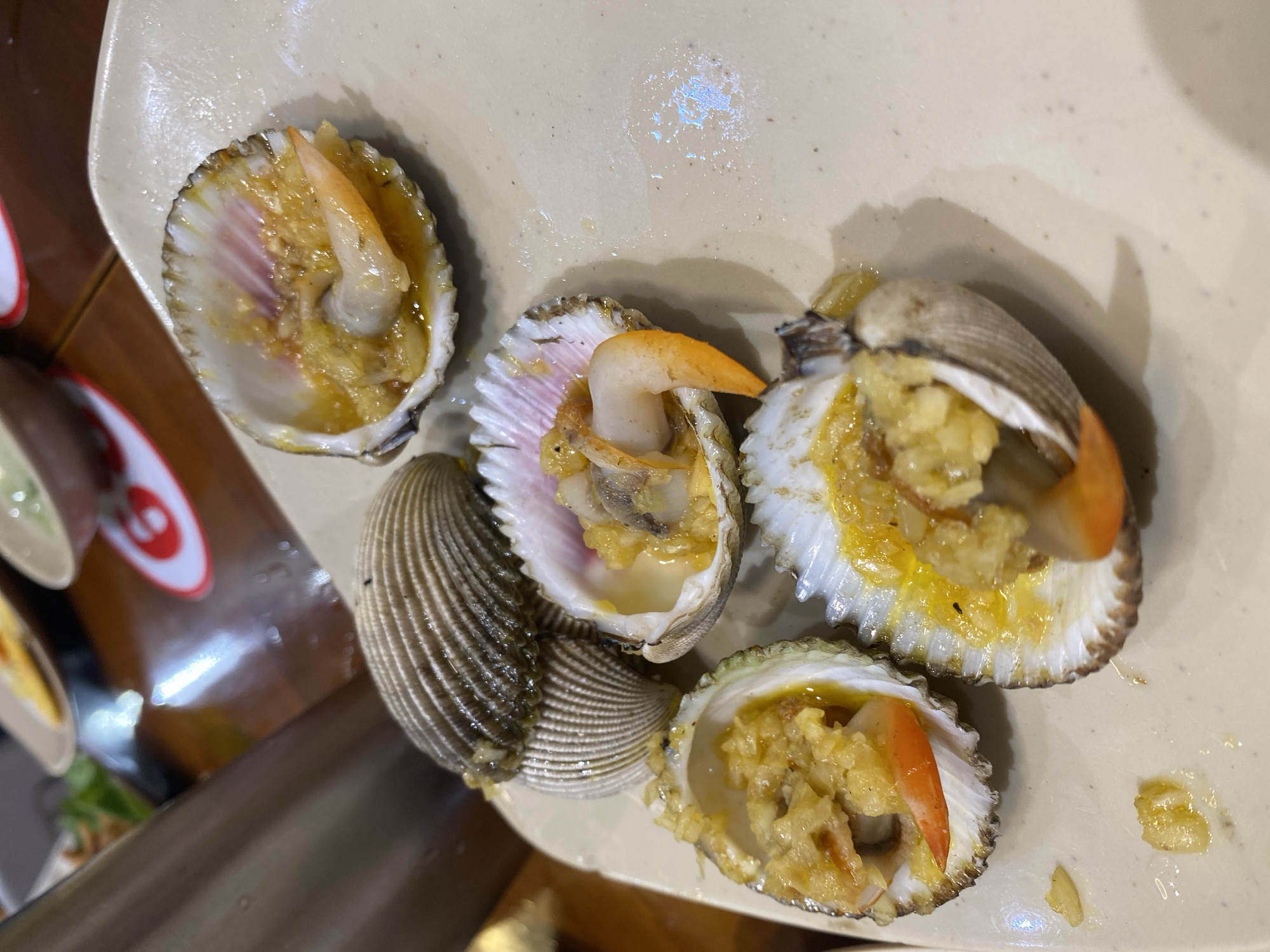 A plate of steamed cockles with garlic and chili is presented in a close-up, slightly elevated shot. The cockles are arranged on a light-colored, oval plate, with some shells open, revealing the cooked mollusks bathed in a yellowish sauce. Orange-colored pieces, likely from chili or shrimp, are visible among the cockles. Two unopened cockles are stacked on top of each other, showing their ribbed shells. The background is blurred, but hints of a wooden table and other dishes suggest a restaurant setting. The overall lighting is dim, with reflections on the table surface. The image quality is moderate, with some noise and motion blur. The scene is likely in Nha Trang Ward, Vietnam, given the cuisine.