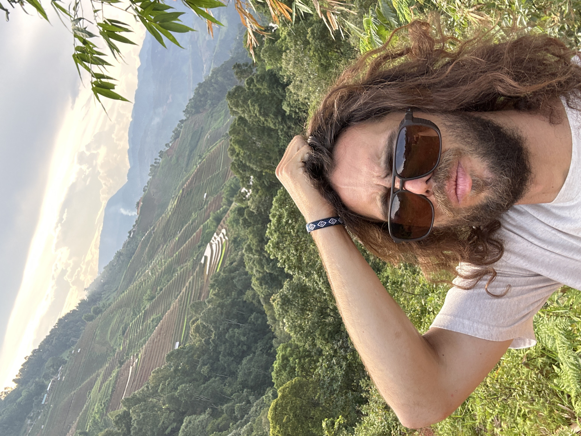 The image is a selfie featuring a man with long, wavy brown hair and a full beard, wearing dark sunglasses and a light-colored t-shirt. He has a blue and white patterned bracelet on his left wrist, and his left hand is casually placed near his forehead, seemingly adjusting his hair. He looks towards the camera with a slight pout.
The photograph is rotated, causing the vast terraced landscape in the background to appear diagonally across the frame. These green, agricultural terraces, characteristic of rice paddies, are carved into rolling hills and stretch far into the distance, indicating a mountainous region known for such cultivation, consistent with Lao Chai, Vietnam. The sky on the left side is bright with soft clouds, suggesting the photo was taken during either early morning or late afternoon, with the sun casting a gentle glow. Lush green foliage and trees cover parts of the hills, and bamboo leaves hang into the shot from the top left corner, adding to the natural setting. The overall mood is one of enjoying the scenic beauty of the outdoors. No text is visible in the image.