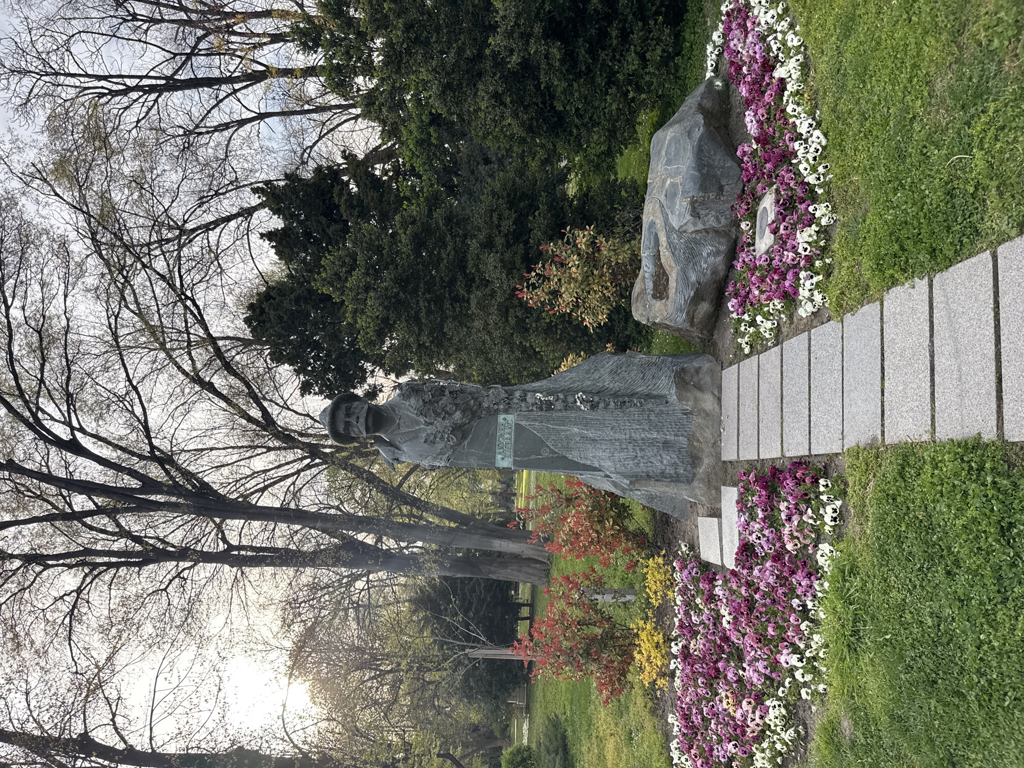 The image shows a statue of Pencho Slaveykov located in Plovdiv, Bulgaria. The statue stands on a stone base, surrounded by flowers, and next to a stone path. Behind the statue are trees and foliage. The sky is visible through the bare branches of the trees.