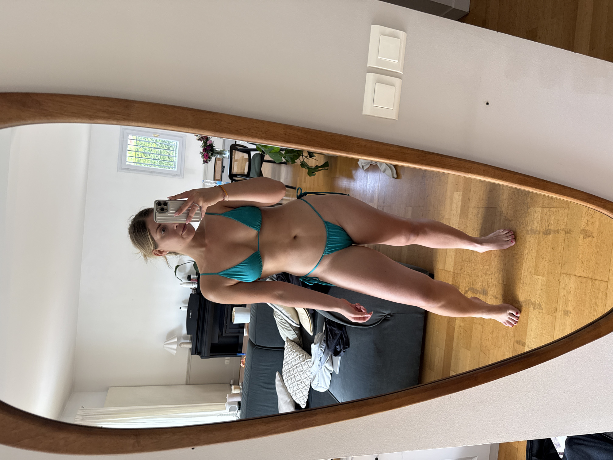 A woman is taking a mirror selfie in a brightly lit room. She is wearing a teal bikini and is standing in front of a large, oval mirror with a wooden frame. Her reflection shows her body from her head to her feet. The room appears to be a living room, with a grey sofa, pillows, and a piano visible in the background. A window with a view of greenery is also present. The floor is made of wooden planks. The image was taken at 10 Rue du Commandant Pilot, Rueil-Malmaison, France.