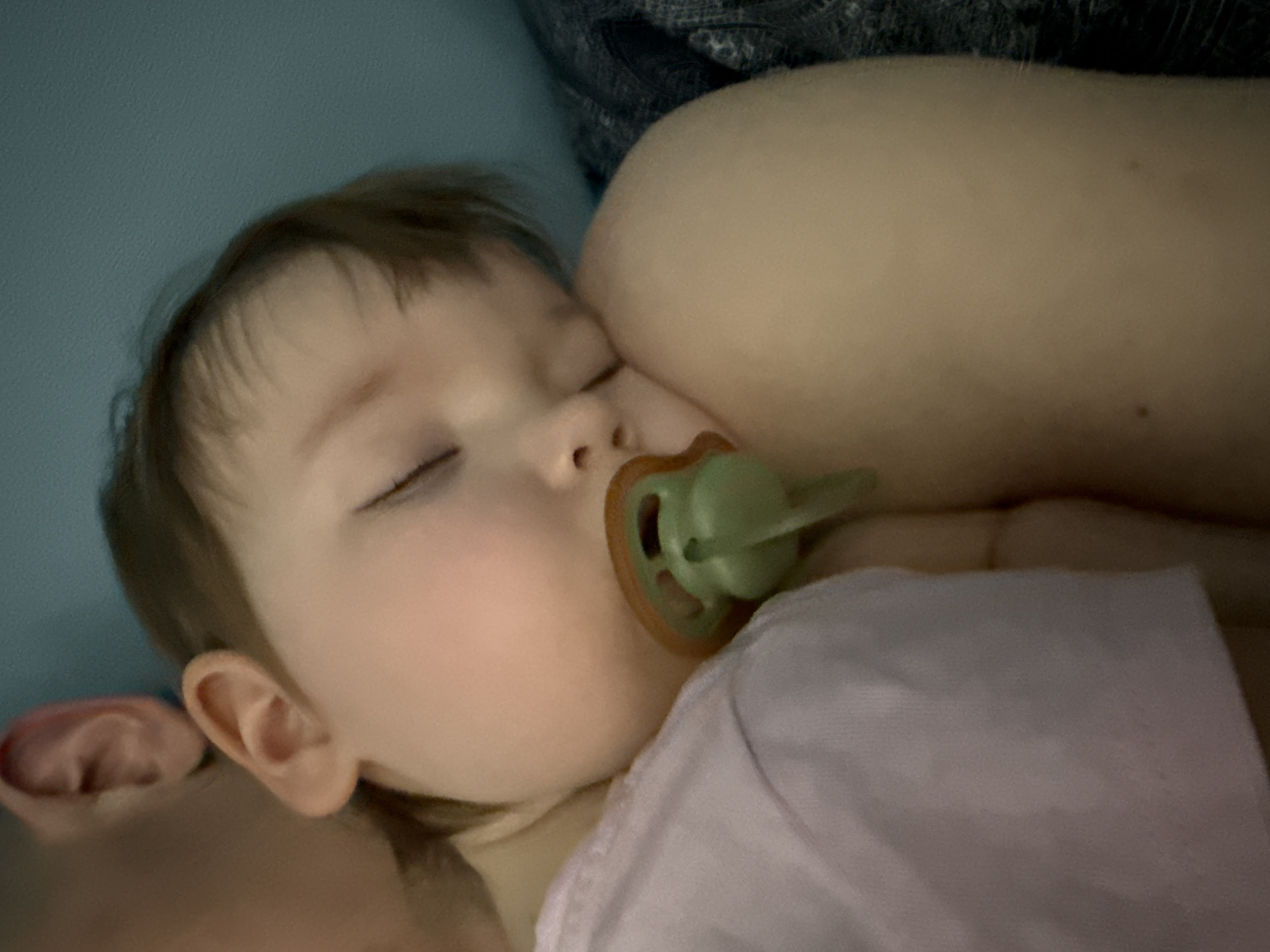 A toddler is asleep, with a pacifier in their mouth, resting their head against a person's chest. The environment consists of bedding, with a blue wall in the background. The location is unspecified and is not discernable as Moscow, Russia.