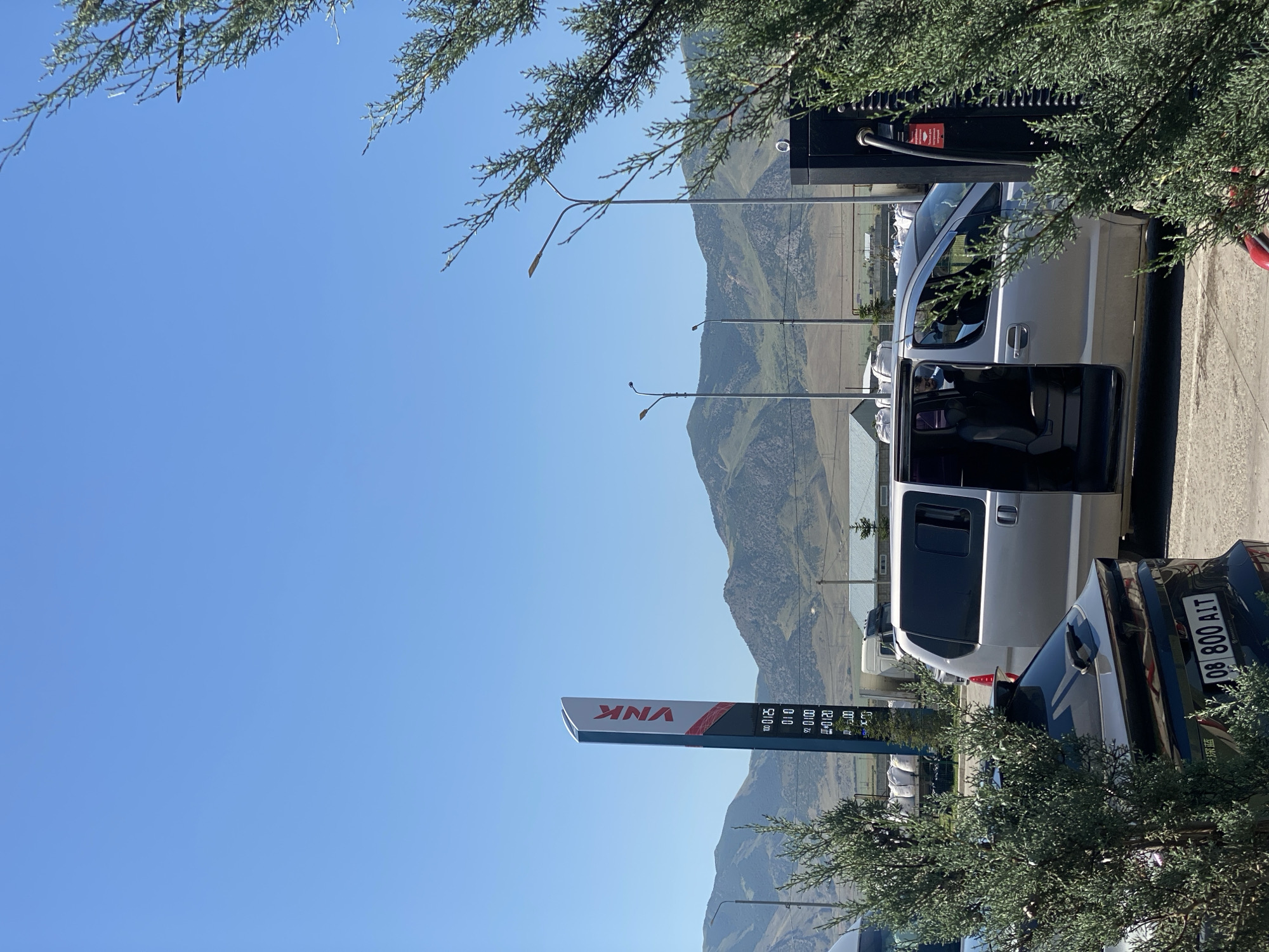 This is a medium shot, eye-level, outdoor photograph taken on a clear day. In the foreground, a dark-colored vehicle with a license plate reading "08 800 AIT" is parked. To its right, a silver minivan with its passenger-side door open is parked next to a black charging station. The minivan appears to have its interior lights on. Behind the vehicles, a tall blue and red sign displaying the word "VNK" and gas prices is visible. In the background, a range of green, treed mountains under a clear blue sky. The environment suggests a rural or semi-rural setting, possibly a gas station or charging facility.