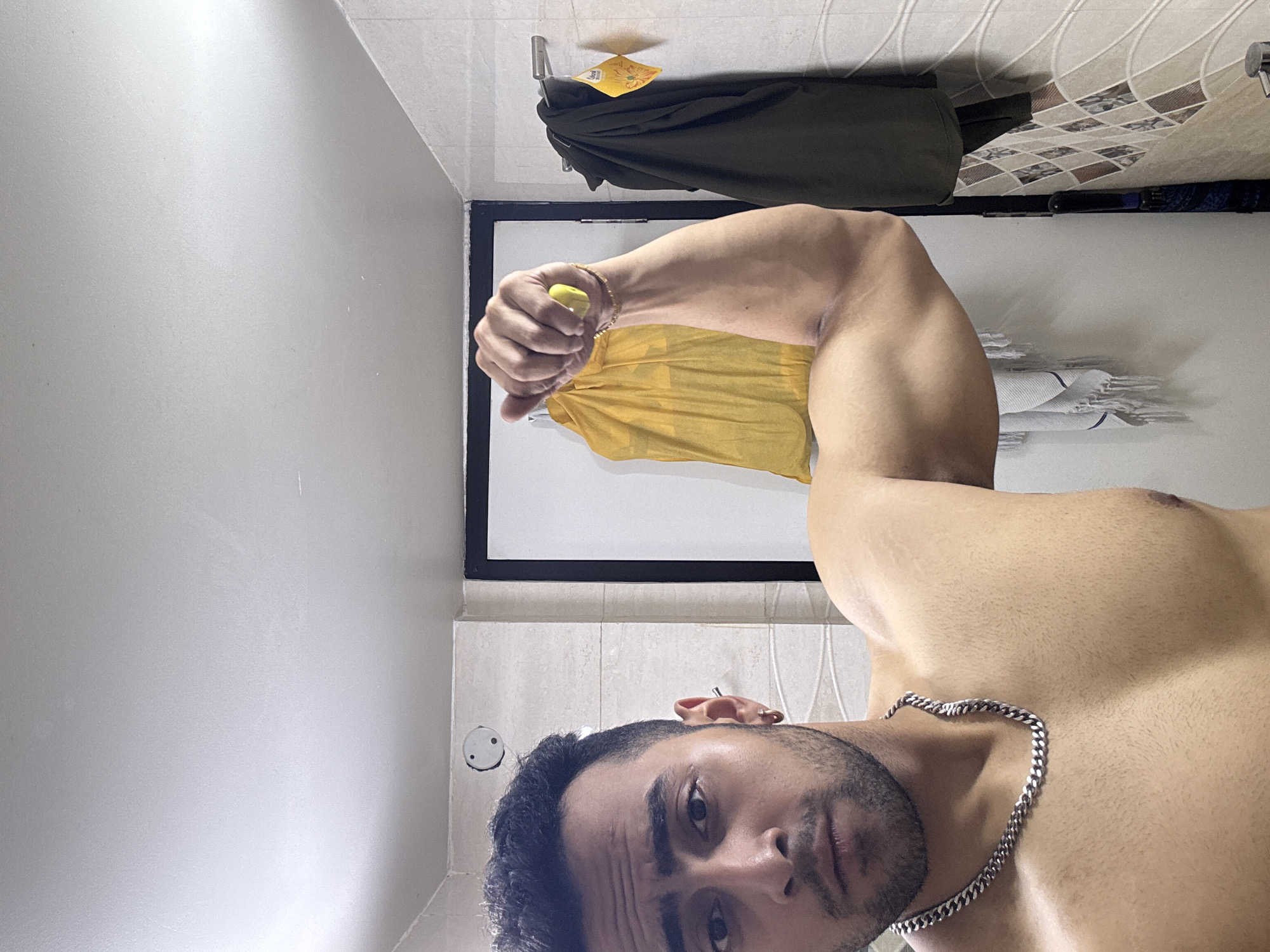 A selfie of a muscular man with a silver chain necklace and an earring in his left ear is taken inside a bathroom. He is flexing his left arm which holds a yellow object in a closed fist. Behind him is a framed mirror in which a yellow garment is reflected. Further behind is a green coat hanging from a hook against a wall with patterned tiles. The man appears to be in his late 20s or early 30s with short, dark hair and facial stubble. The location is Kolkata, India.