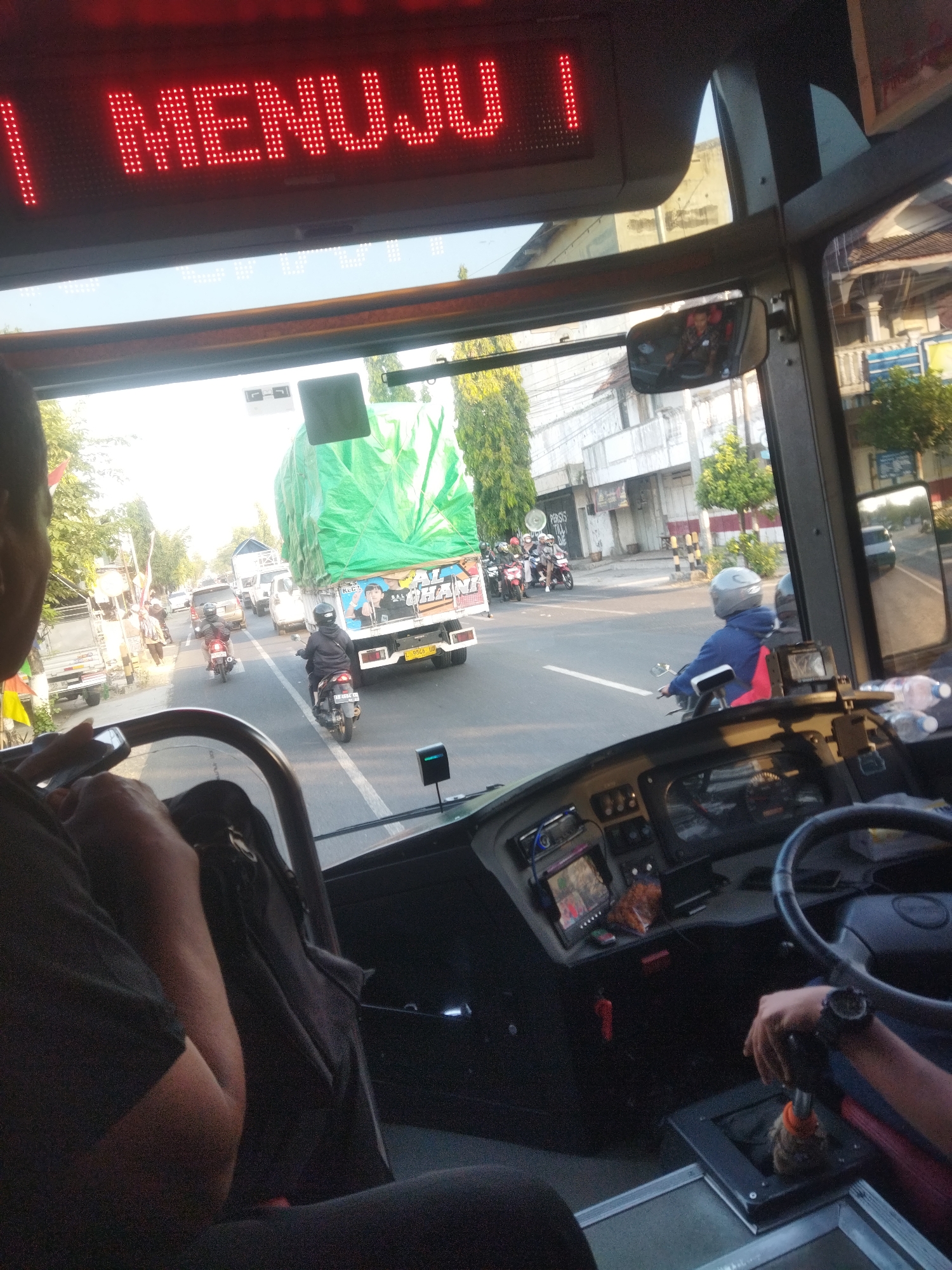 The image is taken from inside a bus, looking forward towards the windshield and the street scene outside. A red LED sign above the windshield displays the text "MENUJU 1". The dashboard of the bus is visible in the foreground, with a steering wheel, gear shift, and various electronic devices. A person's arm and hand are visible on the right side of the frame, resting on the gear shift.
Outside the bus, there is heavy traffic on a street. A large truck with a green tarp covering its load is prominent. Several motorcycles are also on the road, with riders wearing helmets. Buildings and trees line the street, suggesting an urban or semi-urban environment. The time of day appears to be daytime, with sunlight illuminating the scene. The location is identified as Selogiri, Indonesia.