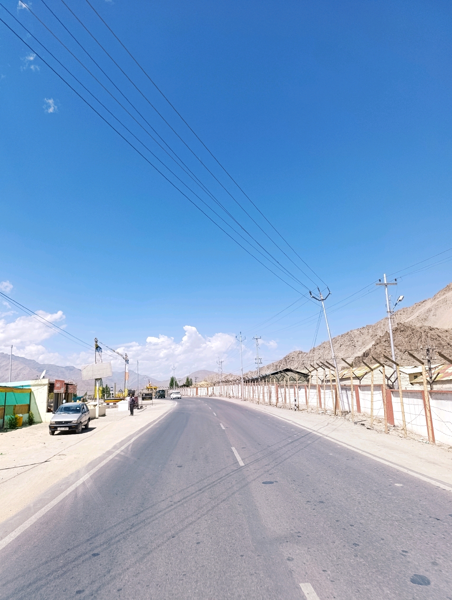 The image shows a wide, paved road in a rural or semi-urban area, likely in the vicinity of Spituk, India, given the context of the provided hints. The road stretches into the distance and is bordered by a sandy shoulder on the left and a fence with barbed wire on the right. Beyond the fence, a barren, mountainous landscape rises, and some buildings are visible. The sky is a clear, vibrant blue with a few scattered white clouds. Several utility poles with wires are present along the roadside, and a few vehicles, including a silver car parked on the left and a white car and another vehicle further down the road, are visible. A single person stands on the roadside near the parked car. The overall impression is one of a sunny day in a dry, high-altitude region.