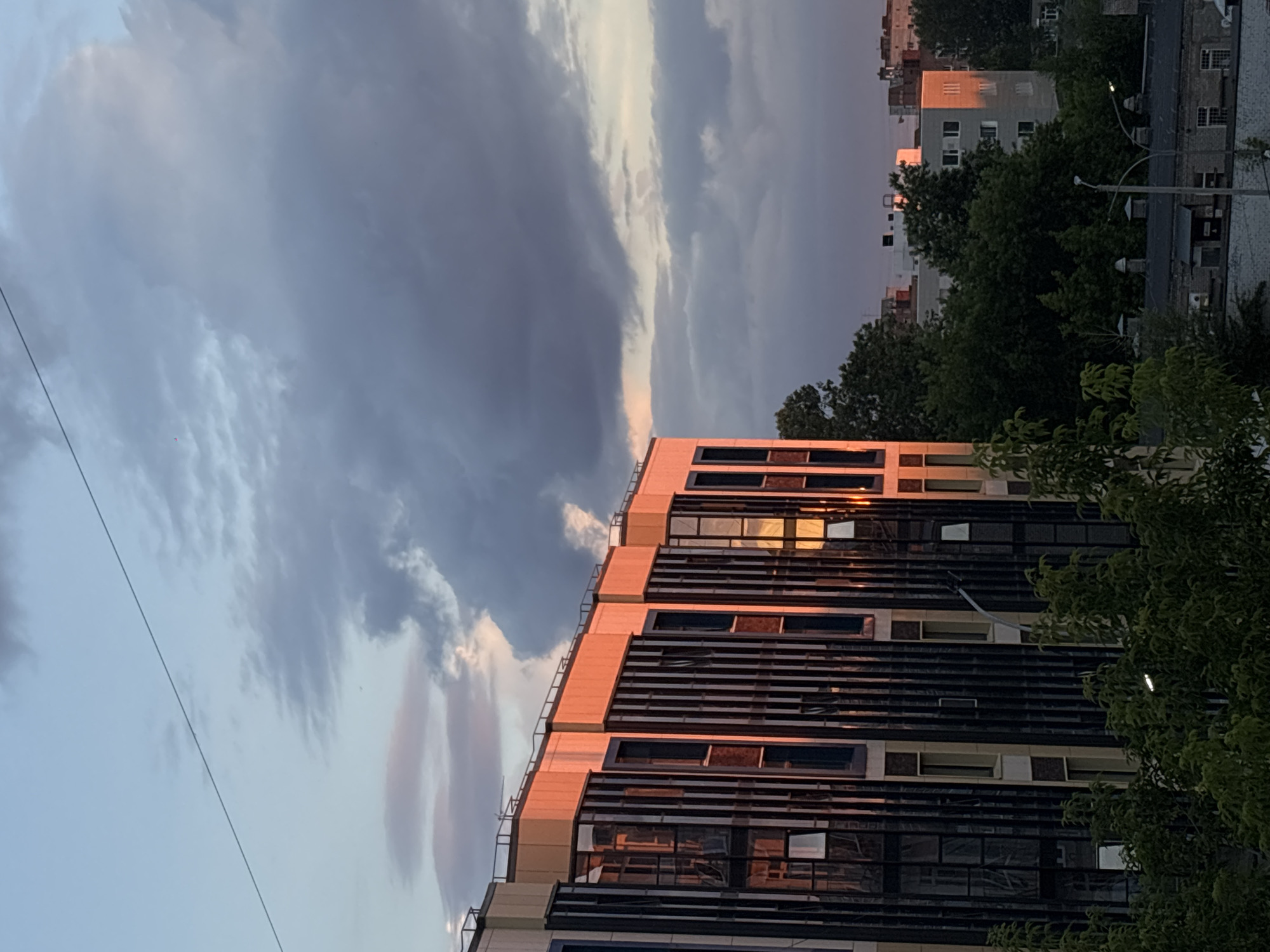 This is a photograph of a city skyline at sunset, likely in Perm, Russia, given the architectural style and the presence of apartment buildings. The foreground features the side of a modern building with a facade of dark glass and lighter colored vertical accents, bathed in the warm, orange glow of the setting sun. Some windows are open, reflecting the sky.
Behind this building, more apartment blocks are visible, some with their sides also catching the sunset light. Dense green trees are scattered throughout the scene, with some in the foreground obscuring parts of the lower floors of the buildings. A power line runs diagonally across the upper portion of the frame against a sky filled with dramatic, dark grey clouds interspersed with softer, lighter clouds and hints of pink and blue from the sunset. In the distance, other buildings and rooftops are visible, some with a slightly older architectural style, suggesting a mix of development within the city. The overall impression is one of an urban environment at the end of the day, with natural light creating a striking visual contrast with the man-made structures and the gathering clouds.