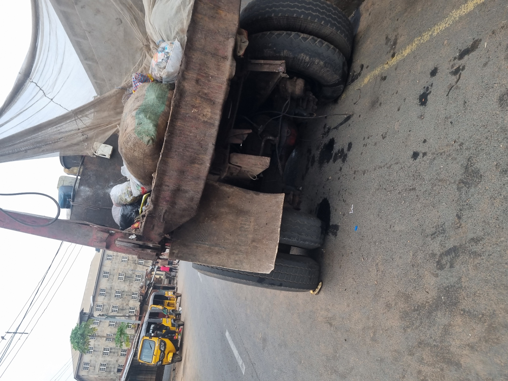 The image captures the rear portion of a heavily laden utility truck, possibly a refuse or recycling vehicle, on an urban street. The truck's body appears old, rusty, and worn, with multiple tires visible at the rear. It is piled high with bags of what appears to be waste or recyclable materials, including a large white bag and a green-patched sack, all partially covered by a large, light-colored mesh or netting.
The truck is positioned on an asphalt road, which shows signs of wear, including oil stains and debris. Faint yellow and white road markings are visible on the tarmac. In the background, to the left of the truck, stand multi-story buildings with numerous small, gridded windows, suggesting an older commercial or residential area. Several yellow three-wheeled auto-rickshaws, commonly known as Keke Napep in Nigeria, are parked or moving on the street alongside the buildings. Utility poles and overhead lines are also visible against a bright, possibly overcast, sky.
The scene depicts a typical busy daytime street in a Nigerian city like Onitsha, characterized by the specific type of public transport vehicles and the general urban environment. No direct human figures are clearly visible, but the setting suggests active daily life. There is no discernible text visible in the image.