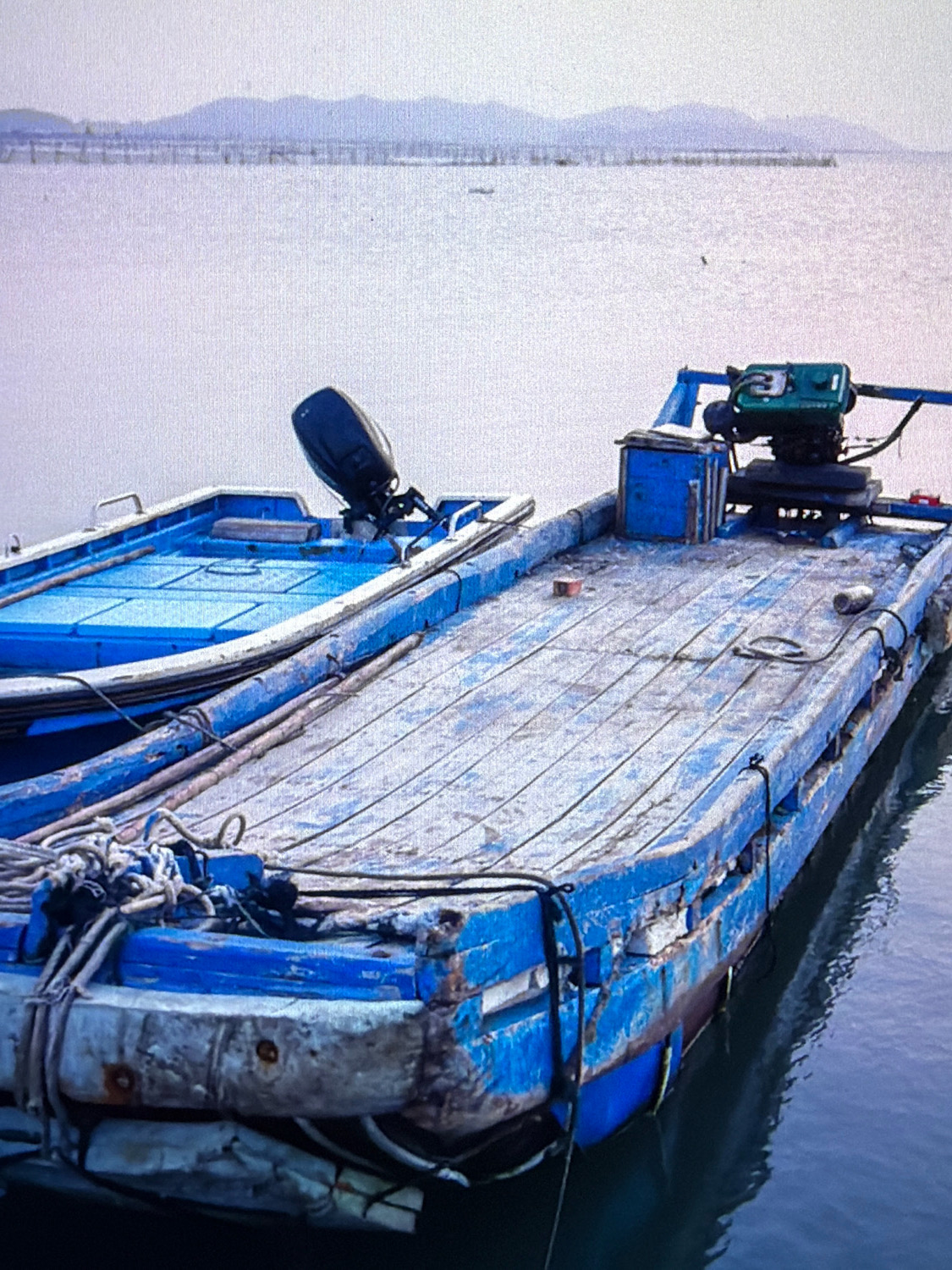 The image shows two boats docked on the water. The foreground boat is a flat-bottomed, weathered wooden barge painted blue with visible signs of wear and tear. It is equipped with a green engine mounted on a platform towards the rear, and several blue crates. Ropes are secured to the barge. A smaller, brighter blue boat with an outboard motor is partially visible to the left, nestled alongside the barge. The water is calm, reflecting the overcast sky. In the background, a body of water extends to a distant shoreline with hazy mountains and what appear to be structures or nets along the horizon. The scene is devoid of people and any specific landmarks that would definitively place it in Kafr Yasif, Israel.