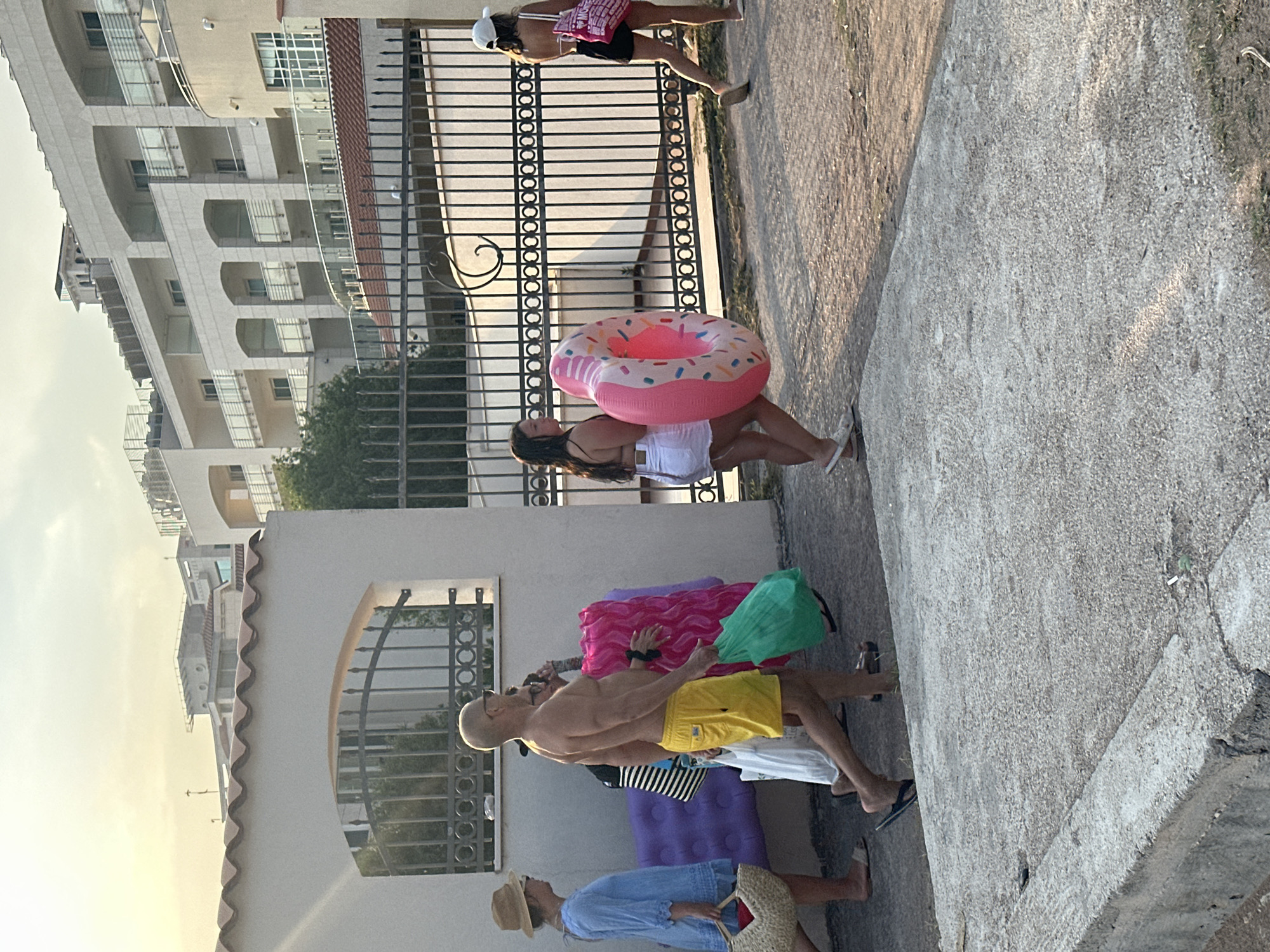 In Boreti, Montenegro, a group of individuals are walking past a fence and a building. The scene is outdoors, with a pathway in the foreground and a residential building with balconies in the background. The sky is visible, with some clouds. A man in yellow shorts is carrying a green bag. A woman in a blue dress is carrying a straw bag. Another woman is carrying a large pink inflatable donut. Another person is walking behind her. The fence appears to be made of metal. The overall impression is of people on holiday.