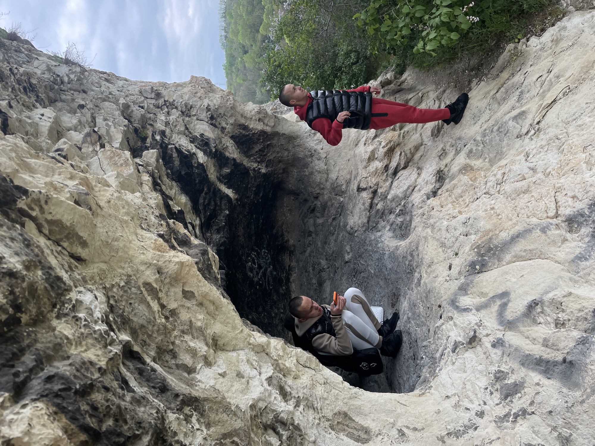 Two men are positioned within a rock cave. The man at the bottom of the frame is seated and looking at an orange smartphone. He is wearing a beige and white sweatsuit, black shoes, and a black vest. The man at the top of the frame is standing. He is wearing a red sweatsuit, black shoes, and a black vest. The cave is primarily light gray rock with areas of dark staining. Green foliage is visible at the top of the frame. The location and city are unknown.