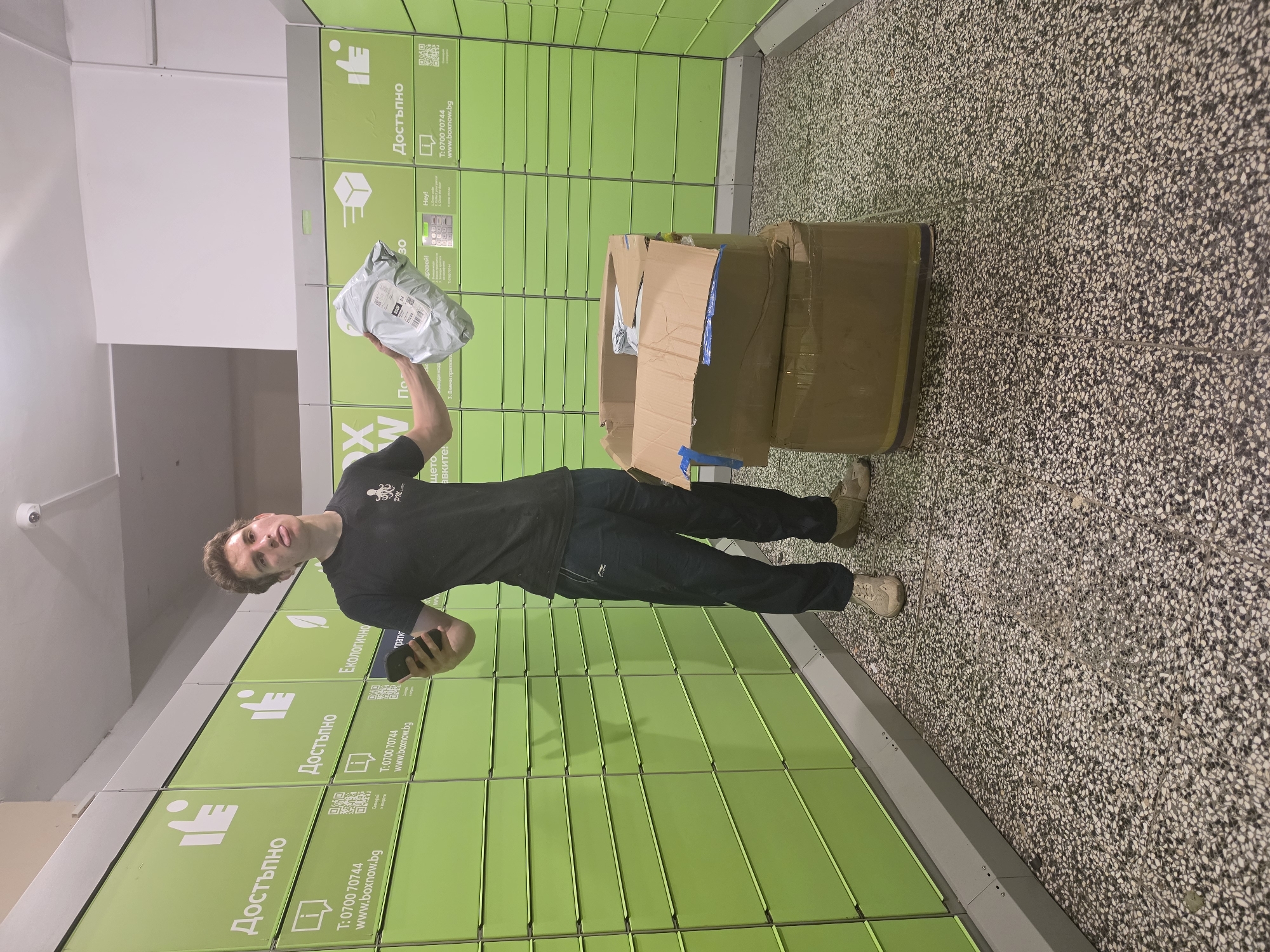 A young man with light brown hair, wearing a black t-shirt and dark athletic pants, stands in front of a row of green parcel lockers in what appears to be a public building in Sofia, Bulgaria. He is holding a smartphone in his left hand and a small white package in his right hand. His facial expression is one of playful exasperation, with his tongue sticking out. To his right, two large cardboard boxes are stacked on the floor. The parcel lockers are labeled in Bulgarian and feature icons indicating "Available" and "Ecological." The floor is a speckled terrazzo. The overall scene suggests the man is in the process of using or interacting with the parcel locker system.