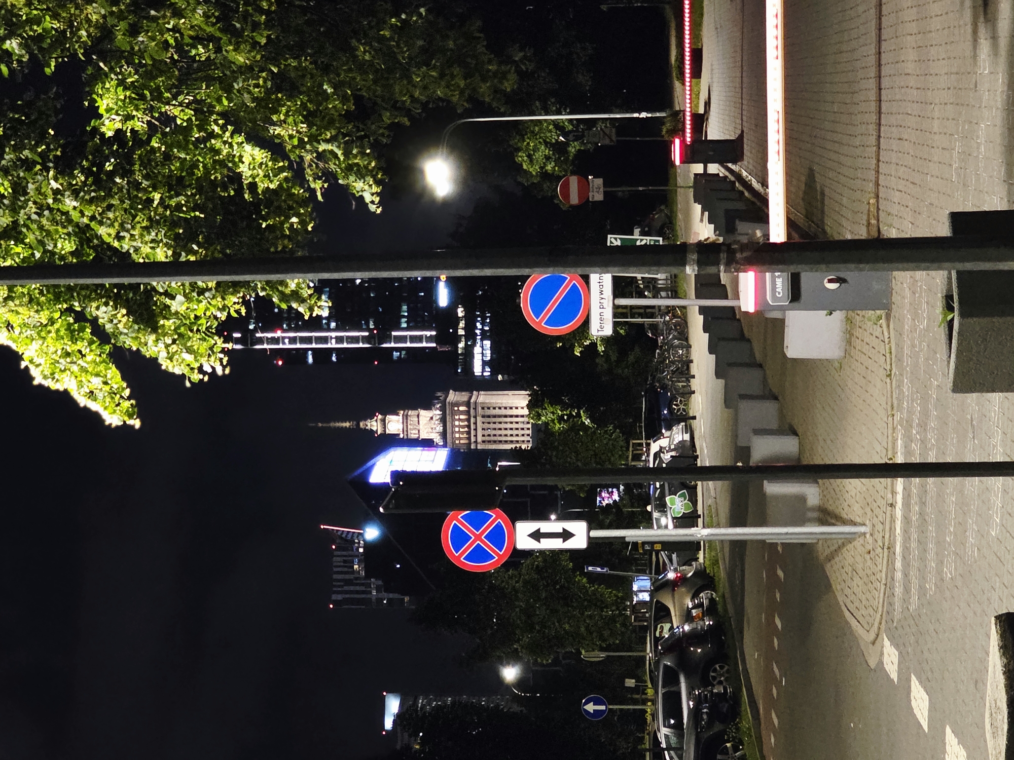 The image depicts a nighttime scene in Warsaw, Poland, likely near the Palace of Culture and Science, which is visible in the background as a prominent landmark. The foreground features a street or parking lot paved with interlocking bricks. Several cars are parked on the left side of the image. Traffic signs are present, including a "no parking" sign (red circle with a blue background and a red X) and a sign indicating two-way traffic. A street lamp illuminates the scene. To the right, a barrier gate system with red illuminated strips is visible, suggesting an entrance or exit to a parking area or controlled zone. The overall environment is urban, with trees bordering the street. The lighting is a mix of streetlights, building lights, and natural illumination from the trees.