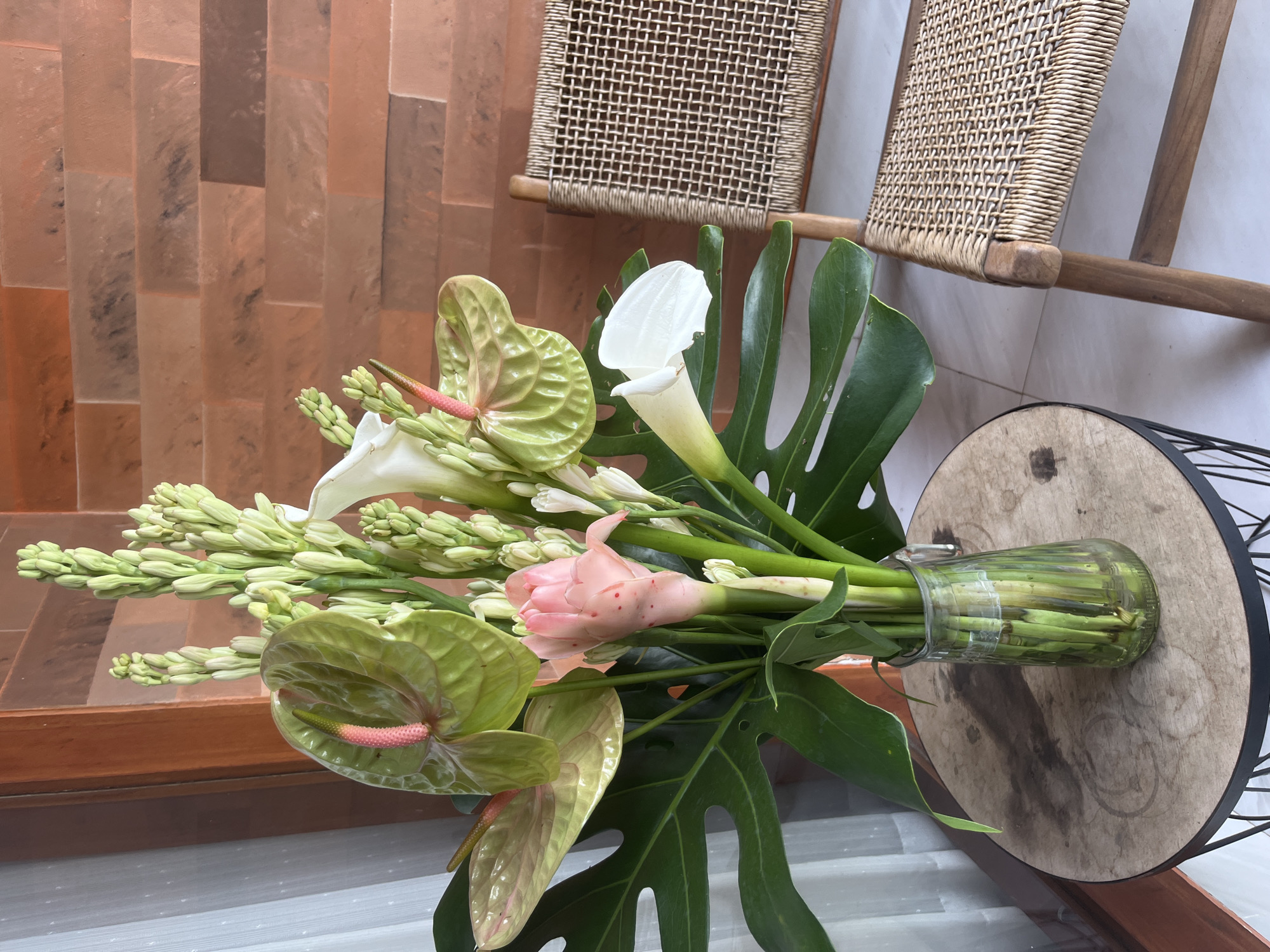 The image shows a floral arrangement in a clear glass vase, consisting of green anthuriums with pink spadices, white calla lilies, unopened white tuberose flowers, a pink ginger flower, and a monstera leaf. The arrangement sits on a round, dark-rimmed table with a wooden top. Behind the table are two woven rattan chairs. The background includes a tiled wall with varying shades of brown and tan. The image was taken in Mengwi, Indonesia.