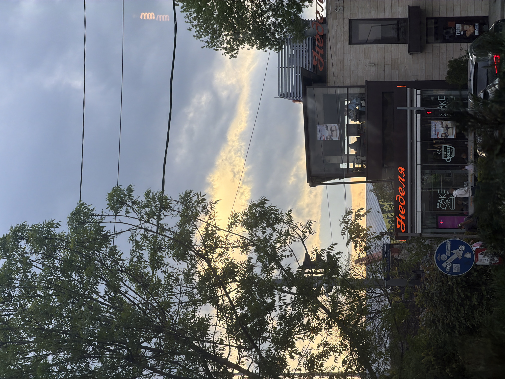 This image captures an urban street scene in Sofia, Bulgaria, during what appears to be either sunrise or sunset, given the warm, golden light streaking through the clouds. The sky is partially overcast but bright where the sun's rays penetrate. Overhead power lines are visible across the upper portion of the frame.
A large, leafy tree with green foliage dominates the left and lower parts of the image, with its branches silhouetted against the bright sky. On the right, a modern building houses a cafe or bakery named "Неделя" (Nedelya), prominently displayed in orange neon letters. Through the cafe's large glass windows, the silhouettes of people can be seen inside, suggesting ongoing activity. Additional text on the cafe exterior includes "CAKE," "FRESH COFFEE & CAKE," and "ГРАБНИ Вкъщи" (Grab it home/Takeaway). The upper facade of the building features stone cladding and a rectangular window.
In the mid-ground, a street sign is visible, partially obscured by foliage, displaying "Акад. ... Стефанов" (Acad. ... Stefanov), which is a street name. A blue circular road sign with white pictograms is also present near the cafe entrance. The rear of a dark-colored car is seen on the far right. A peculiar visual detail in the sky are light reflections resembling "mmm" letters. The overall scene depicts a typical urban environment with shops and street infrastructure, bathed in the soft light of dawn or dusk.