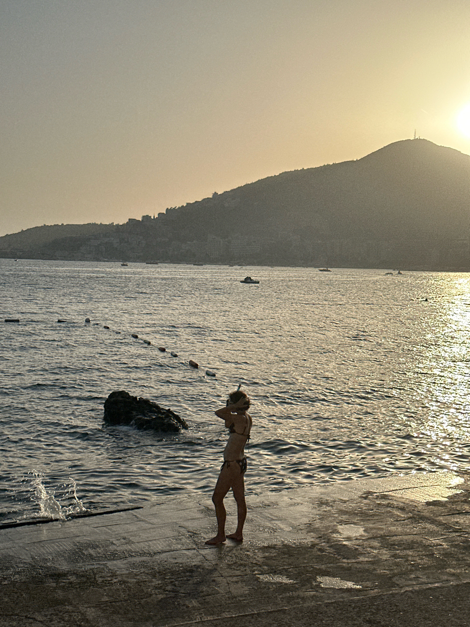 A full-body, outdoor, medium shot captures a person in a bikini and snorkel gear standing on a paved waterfront area at sunset. The person is silhouetted against the water, facing away from the camera and towards a hazy, sunlit mountain range in the background. The water is calm with gentle ripples, and sunlight reflects off its surface. A line of buoys stretches across the water in the middle ground, and a large rock is visible near the shore to the left. Waves crash against the paved area on the left, creating a spray. The sky is a soft gradient of yellow and gray. The scene appears to be the Budva waterfront in Montenegro.
