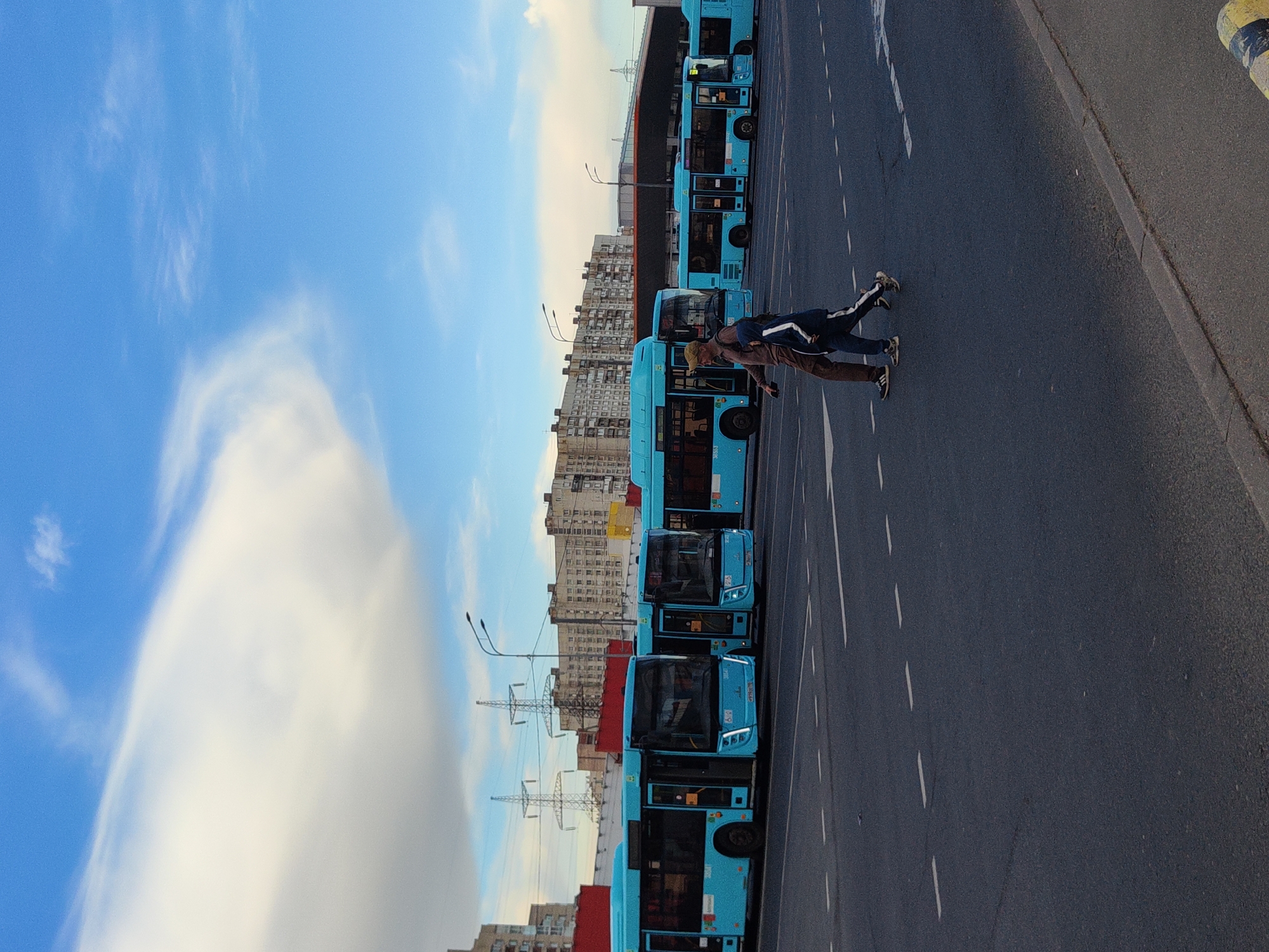 In this image, two people are seen from a high angle, appearing to be walking or running on a road. They are dressed casually in athletic wear. The scene is an urban setting, likely in Saint Petersburg, Russia, as suggested by the architecture of the buildings in the background. A line of blue buses is parked along the side of the road, suggesting a bus depot or waiting area. The sky is blue with scattered white clouds, indicating fair weather. The overall impression is of an ordinary day in a city, with the people engaged in some form of physical activity. The image is oriented vertically, with the road and buses running down the right side of the frame and the sky and buildings to the left. The time of day appears to be daytime, possibly late afternoon given the lighting. There are no discernible emotions visible from the distant figures. There is no visible text in the image.
