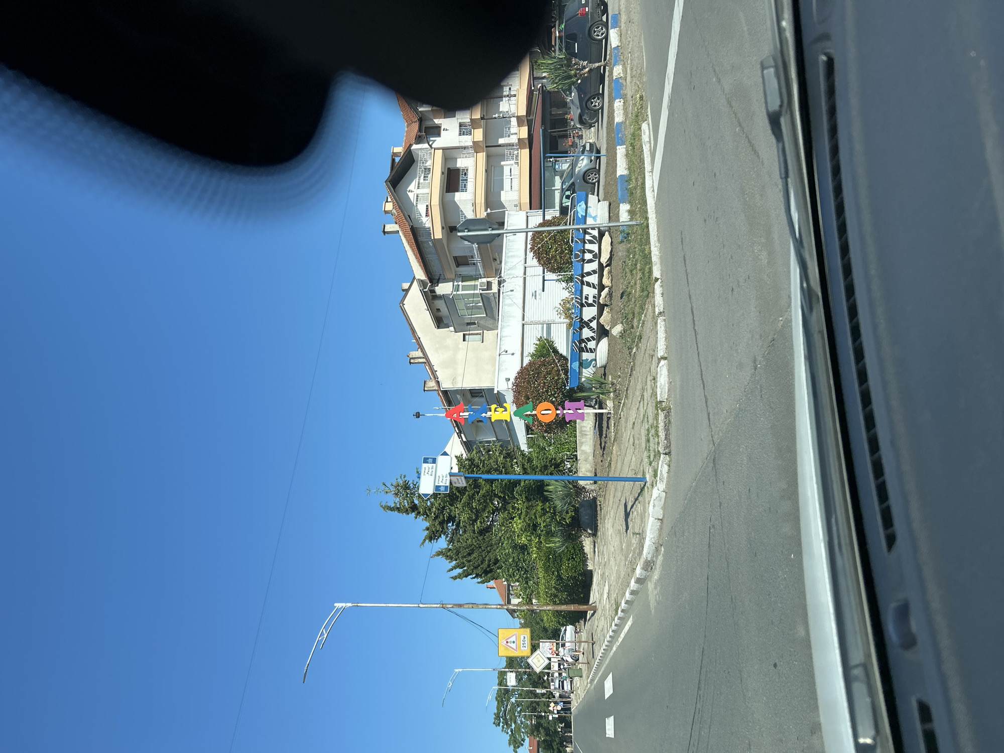 The image depicts a street scene in Aheloy, Bulgaria, on a clear, sunny day. The viewpoint is from inside a vehicle, looking forward and slightly to the right through the windshield. In the foreground, the dashboard and windshield wipers of the vehicle are visible.
The street itself is paved, with white lane markings and a sidewalk lined with a white curb. On the sidewalk, a blue pole with street signs is prominently positioned. These signs appear to be directional, with text that is difficult to read clearly due to distance and resolution. Adjacent to the pole, large, colorful, three-dimensional letters spell out "AKHELOI" vertically. The letters are red, orange, yellow, green, and purple, mounted on a white base with blue trim. Behind the letters, a low wall with blue and white trim encloses a small area of vegetation.
Further down the street, a row of trees and bushes line the sidewalk. Beyond the vegetation, a multi-story building with light-colored walls and red-tiled roofs is visible. The building has balconies and windows. Several cars are parked on the side of the street, including a silver car and a darker colored SUV. In the distance, more buildings and trees are visible under a clear blue sky. A street lamp with a modern design stands on the left side of the road. A yellow triangular traffic sign with a red border and a graphic of a car is also visible on the left. The overall impression is of a coastal town or a town on a sunny day, with a mix of residential and commercial buildings.