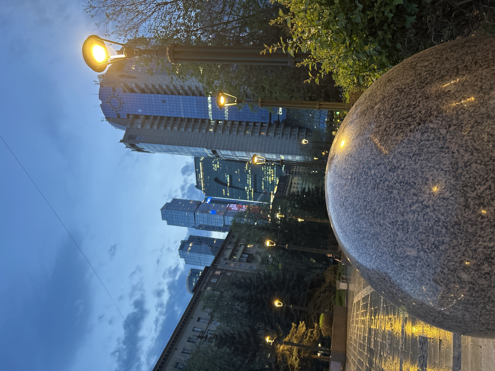 A ground-level, medium shot, taken at dusk or twilight, is composed of the stone round object in the foreground, row of street lights on the side of the path, high rise buildings, and sky. The round object is of a mottled gray shade and shows the reflection of street lights. The street lights line the brick-paved path and stretch back into the middle ground. Behind the street lights are some tall buildings with lighted windows. Buildings are constructed of glass, metal, and brick. The sky is blue, with gray clouds in the lower left of the scene. Tall trees with green foliage can be seen in the right side of the scene.