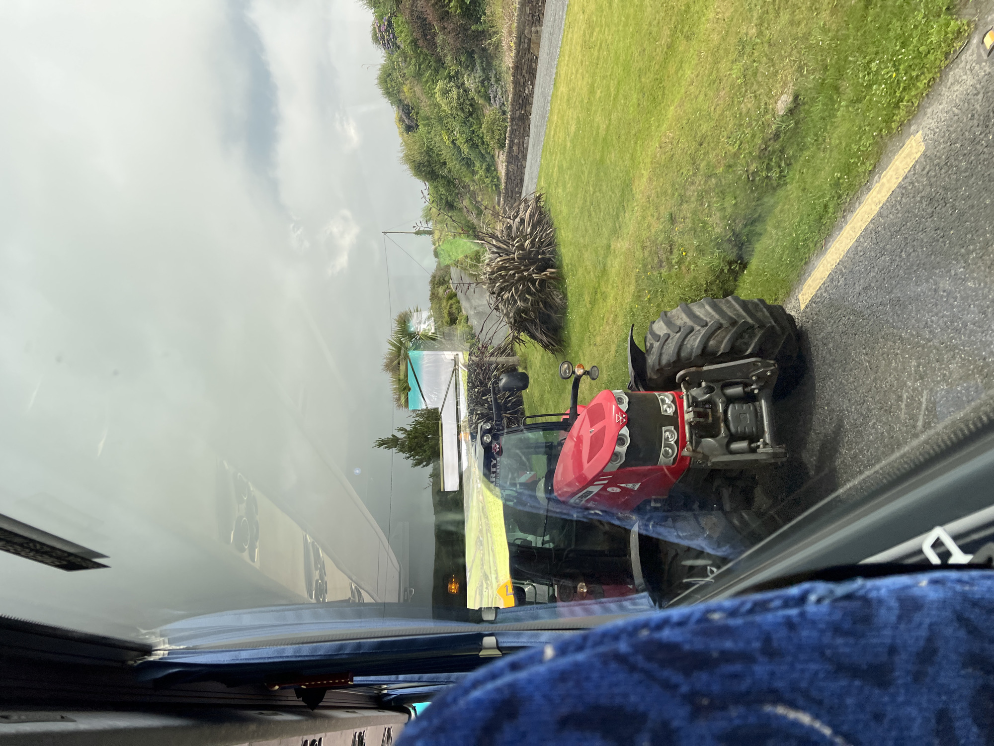 The image, taken from inside a vehicle, displays a red Massey Ferguson tractor parked on the side of a road in Dunfanaghy, Ireland. The tractor is the central focus, positioned parallel to the road with its front facing towards the viewer. A yellow line is visible on the asphalt road, separating the lane from the grassy verge. The grassy area is lush and green, with scattered white flowers. Beyond the grass, a stone wall runs along the edge of a property, and a gravel path leads upwards. The landscape is characterized by rolling hills covered in dense, green vegetation, with some scattered trees and bushes. The sky is overcast with diffused light. Reflecting on the vehicle's window, parts of the interior, including a blue seat, are visible.