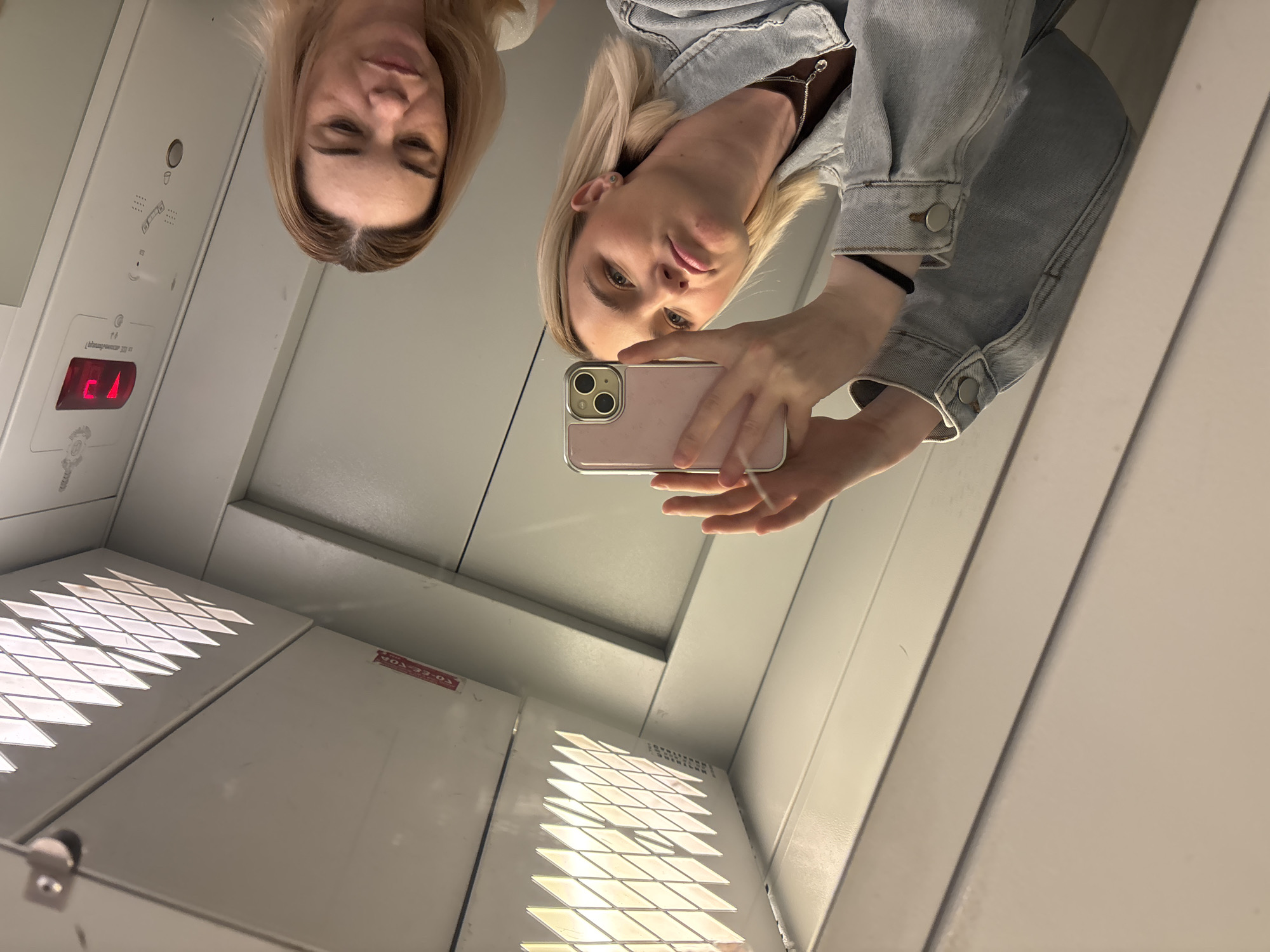 Two fair-skinned women are in an elevator, likely in Vsevolozhsk, Russia, given the provided context. One woman, wearing a light blue denim jacket, is holding a pink iPhone and taking a selfie. The other woman is standing beside her. The elevator interior is white with a lighted display showing "C." The elevator's ceiling has a vent with diamond-shaped openings.