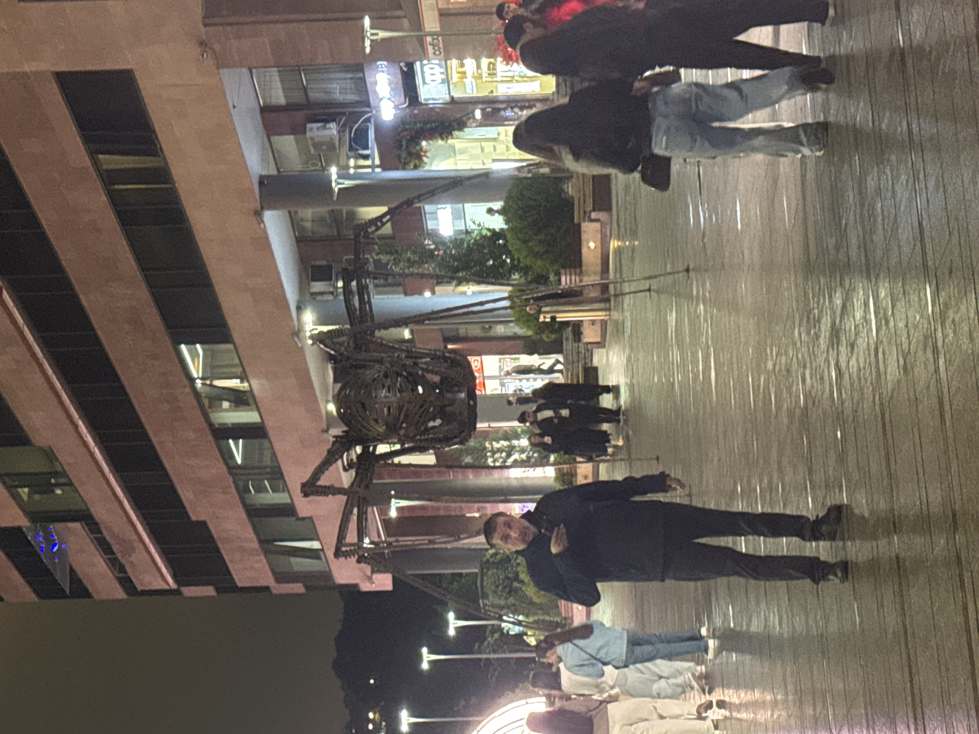 The image depicts a night scene in Yerevan, Armenia, on a pedestrian walkway. The image includes a large, metal abstract sculpture, possibly a dragon or mythological creature. A tall, modern building with rows of windows is on the left. Several people are present, some walking in groups. A man in a dark jacket and pants is standing and looking towards the camera. The pedestrian area appears to have wood-like flooring and is illuminated by street lights. In the background, there are buildings with shops and restaurant signs in Armenian.