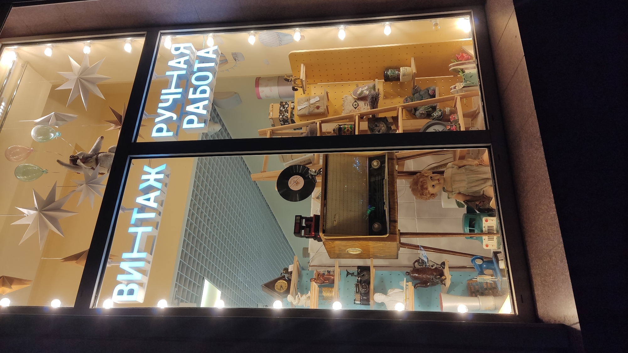 The media file shows a nighttime view of a shop window display in Krasnodar, Russia. The window is divided into two sections by a dark frame. The left section features a display of paper stars, balloons, and what appears to be a stuffed animal or puppet. The right section showcases shelves filled with various items, including jars, boxes, and what looks like a vintage radio.
Prominently displayed in the window are two illuminated signs with Cyrillic text. The sign on the left reads "ВИНТАЖ" (VINTAGE) in large, blue neon letters. To the right of this, also in blue neon, is the text "РУЧНАЯ РАБОТА" (HANDMADE).
Beneath the "ВИНТАЖ" sign, within the window display, are more shelves and objects. A vintage-style radio is visible, along with a vinyl record, a clock, and several figurines or decorative items. In the lower part of the right section of the window, a doll with blonde curly hair is seated, and below it are various tools and objects.
The overall scene suggests a retail establishment specializing in vintage and handmade items, with a creative and somewhat eclectic display designed to attract attention at night. The lighting within the display creates a warm and inviting atmosphere.