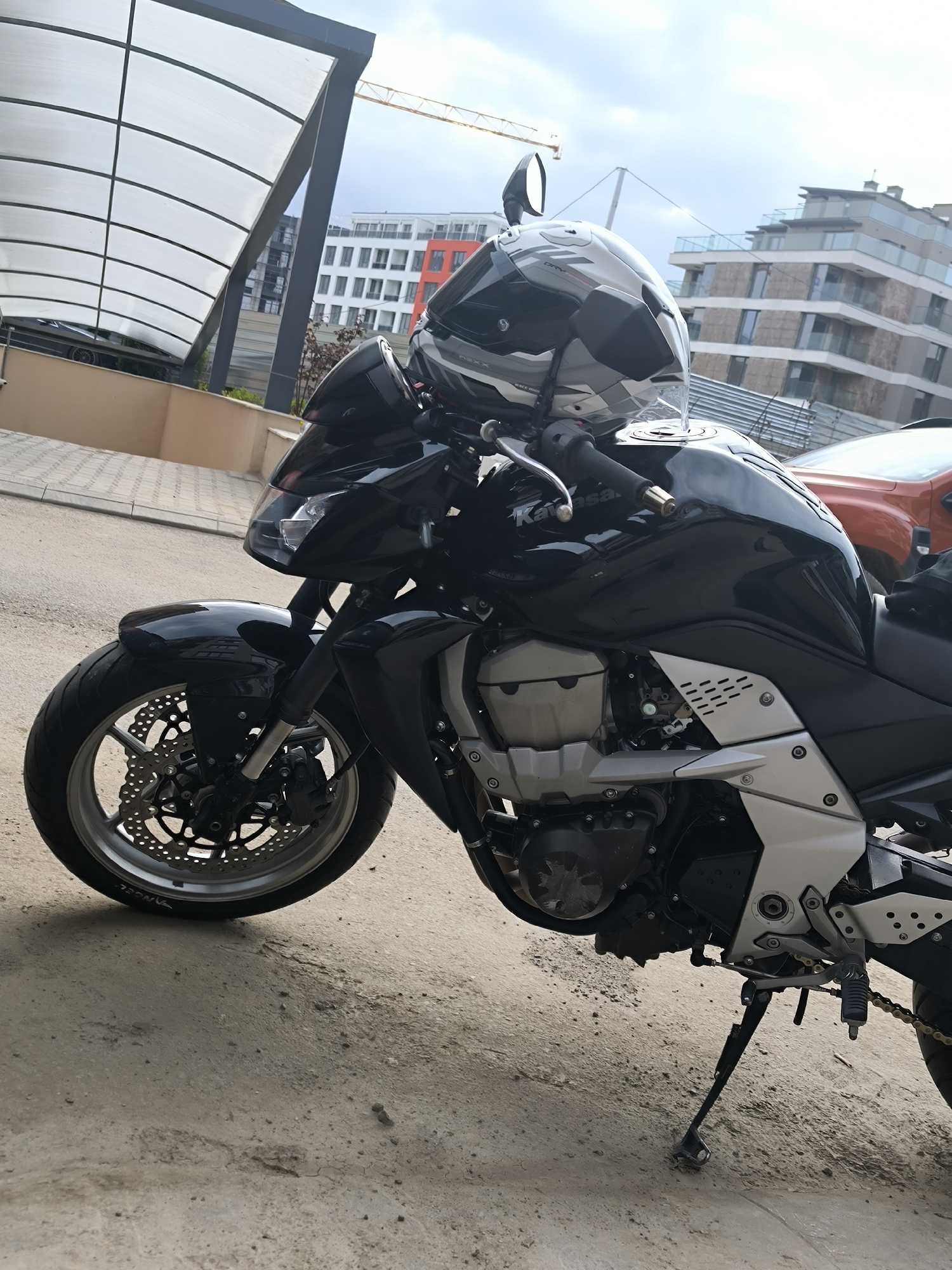 A black Kawasaki motorcycle is parked outdoors in Sofia, Bulgaria, with a grey and black helmet resting on its fuel tank. The motorcycle features a glossy black finish, silver-colored engine components, and metallic frame accents. The front wheel displays disc brakes with perforated rotors, and the word "ANGEL" is visible on the tire sidewall. The kickstand is extended, indicating the motorcycle is stationary.
The scene unfolds in an urban or suburban setting during daytime, under a partly cloudy sky. In the background to the left, a modern building with a distinctive white, translucent arched canopy structure supported by dark grey pillars is visible. Adjacent to it is another multi-story building with white facades and sections featuring red/orange cladding. A yellow construction crane is visible above these structures, suggesting ongoing development. To the right, another contemporary building with multiple glass balconies and railings can be seen, partially obscured by the motorcycle and an orange car.
Visible text includes "Kawasaki" on the motorcycle's fuel tank and "DRY" and "NEXX" on the helmet. The motorcycle appears clean and well-maintained, ready for use.