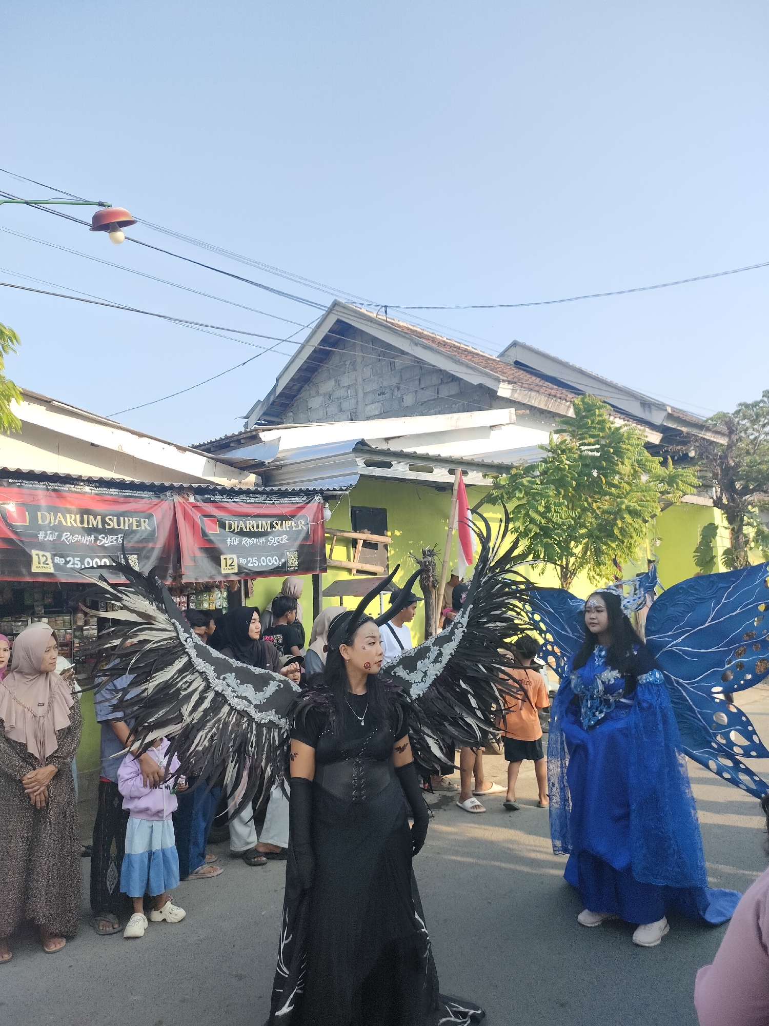 The image depicts a street scene in Tongaswetan, Indonesia, on a clear day with a bright blue sky. In the foreground, two individuals are dressed in elaborate costumes. The person on the left wears a black, floor-length gown with large black and white feathered wings, and black gloves. They have facial markings and dark hair. The person on the right is adorned in a vibrant blue, fairy-like costume with large blue butterfly wings.
A crowd of people is visible in the background, some of whom appear to be spectators. There are banners visible that say "DJARUM SUPER #IniRasanya SUPER" and show a price of "12 RP 25.000," suggesting a commercial event or promotion. Buildings with green walls and light-colored roofs line the street. Overhead, electrical wires and a red street lamp are present. A small Indonesian flag is also visible. The ground is paved, indicating a street or public area. The overall atmosphere suggests a festive occasion or parade.