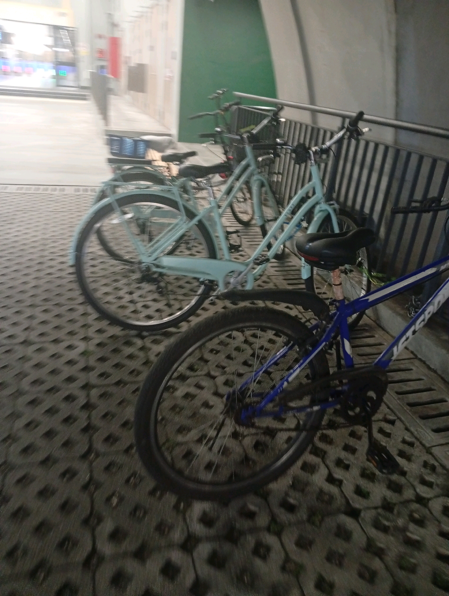 The image shows a bicycle rack with two bicycles parked. One bicycle is light blue and appears to be a cruiser bike, with a basket attached to the front. The other bicycle is blue and white, and appears to be a mountain bike. Both bicycles are parked on a patterned concrete surface, and a metal fence is visible in the background. The scene is outdoors and appears to be in an urban environment. There are no people visible in the image.
