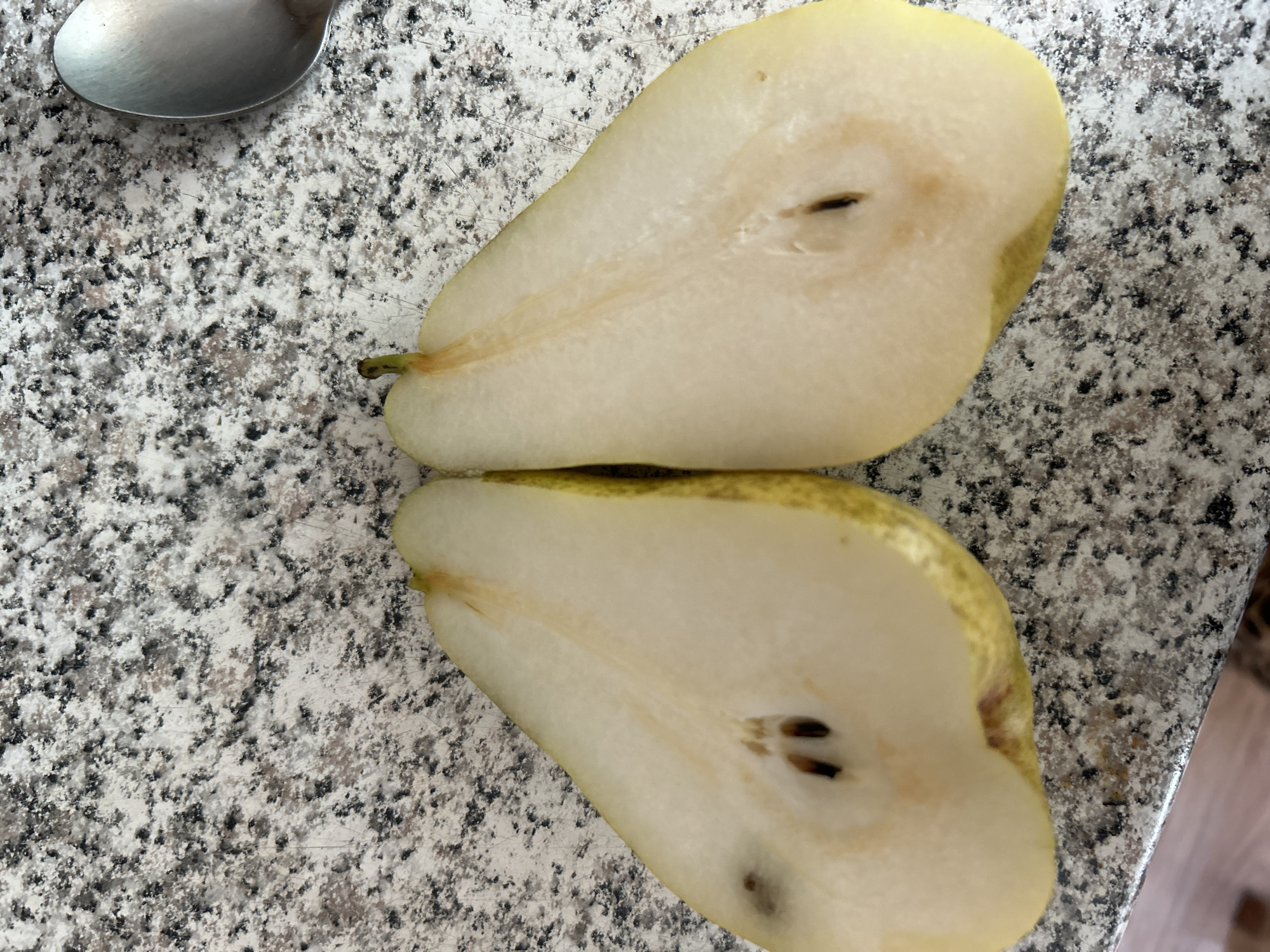 Two halves of a pear are displayed on a speckled granite countertop. The pear halves are positioned side-by-side, with their cut surfaces facing upward, revealing the seeds and the pale yellow flesh. The pear on the left has a few dark spots visible on its surface, while the pear on the right appears unblemished. A portion of a silver spoon is visible in the upper right corner of the image. The environment is a domestic setting, likely a kitchen, given the countertop material and the presence of a spoon. The image does not provide information about the city of Калицино, Russia.