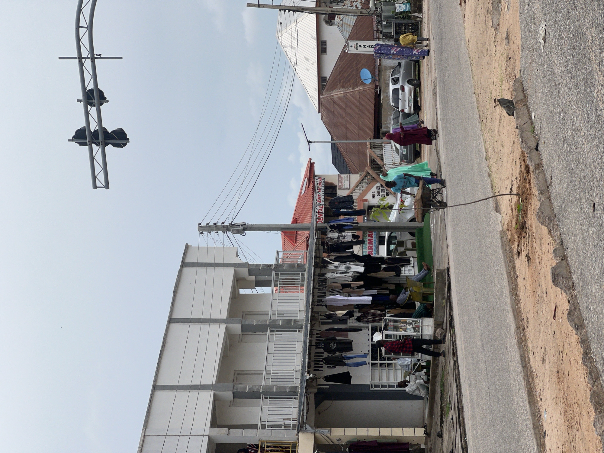 This image depicts a street scene in Jalingo, Nigeria, under an overcast sky. A two-story building with a white facade and gray concrete pillars dominates the left side of the frame. The ground floor houses a shop displaying various clothing items hanging from a rack outside. Several individuals are visible, some sitting on the ground near the shop, while others stand. A person in a red and black checkered shirt walks in front of the shop.
Across the street, a road is visible with a sandy verge on the right. Several vehicles are parked or driving on the road, including a silver sedan, a darker sedan, and a white car. A traffic light is mounted on an overhead structure, though it appears to be covered or out of order. Further down the street, more buildings are present, some with corrugated metal roofs. Two women in brightly colored traditional clothing are walking on the sidewalk, one pushing a wheelbarrow. A young child in a yellow shirt is walking near the end of the street. A bird is perched on a low concrete barrier in the foreground. The scene suggests a busy commercial area with a mix of residential and retail establishments.
