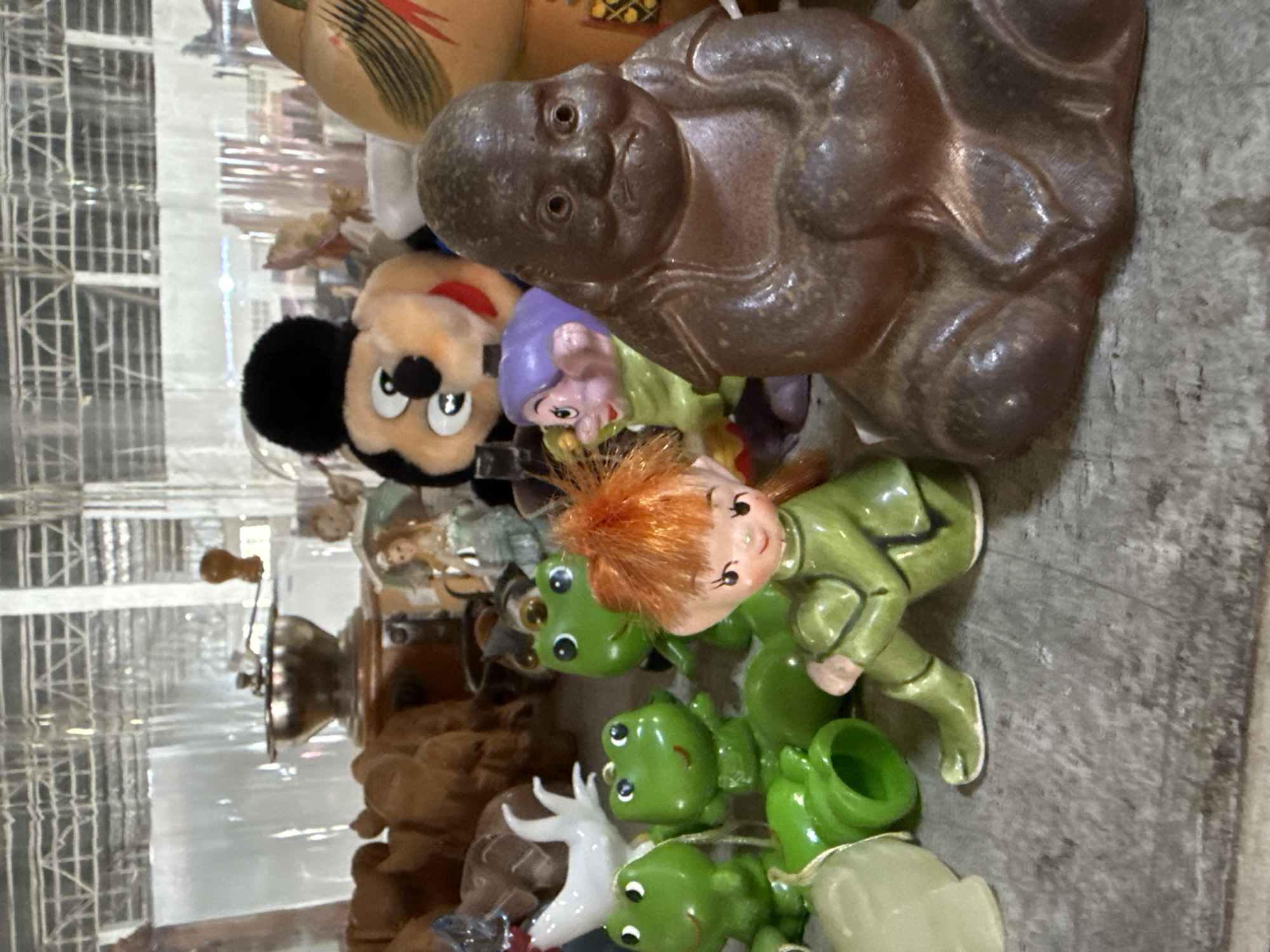 The image shows a collection of decorative figurines and objects displayed on a textured gray surface, possibly a tabletop or shelf. In the foreground, to the right, is a dark brown, seated figurine with a wrinkled face and open eyes, appearing contemplative. To the left of this figurine is a small ceramic figure of a girl with red hair and green clothing, sitting cross-legged. Several green ceramic frog figurines are positioned in front of the girl and further to the left. Behind these figures, a plush Mickey Mouse toy is visible, and to its right, a portion of a gourd-like object with red and black markings. In the background, a vintage coffee grinder and other indistinct objects and translucent partitions can be seen. The overall impression is that of an indoor display of various collectible items. There is no indication of the location being Chalong, Thailand, or any specific city.