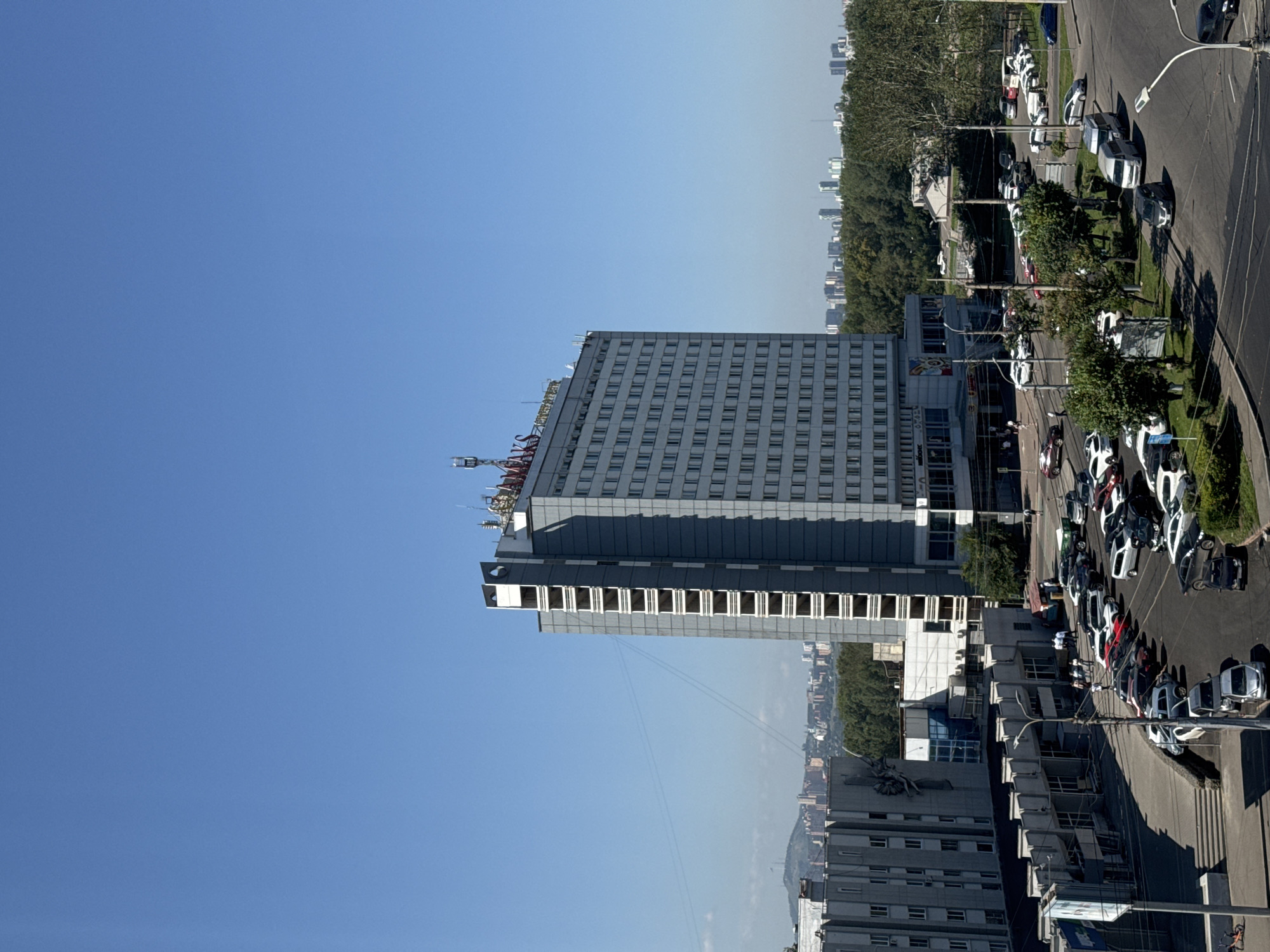 The image captures an urban scene in Krasnoyarsk, Russia, on a clear day. Dominating the frame is a tall, grey, multi-story building, likely a hotel or office building, with numerous windows and some visible scaffolding or unfinished sections on its upper floors. Red lettering spelling "MAKS" is visible at the top of this building. To the left, another, shorter grey building with a similar architectural style is present. In the foreground, a street intersection is visible, with several cars parked and driving on the asphalt. Some of these vehicles are white, grey, and black. A few individuals can be seen walking on the sidewalk and near the buildings. Lush green trees are interspersed throughout the scene, with a dense treeline forming a backdrop to the urban structures. In the distance, more buildings and a hilly landscape are visible under a clear blue sky. The overall impression is one of a functional, somewhat dated urban environment.