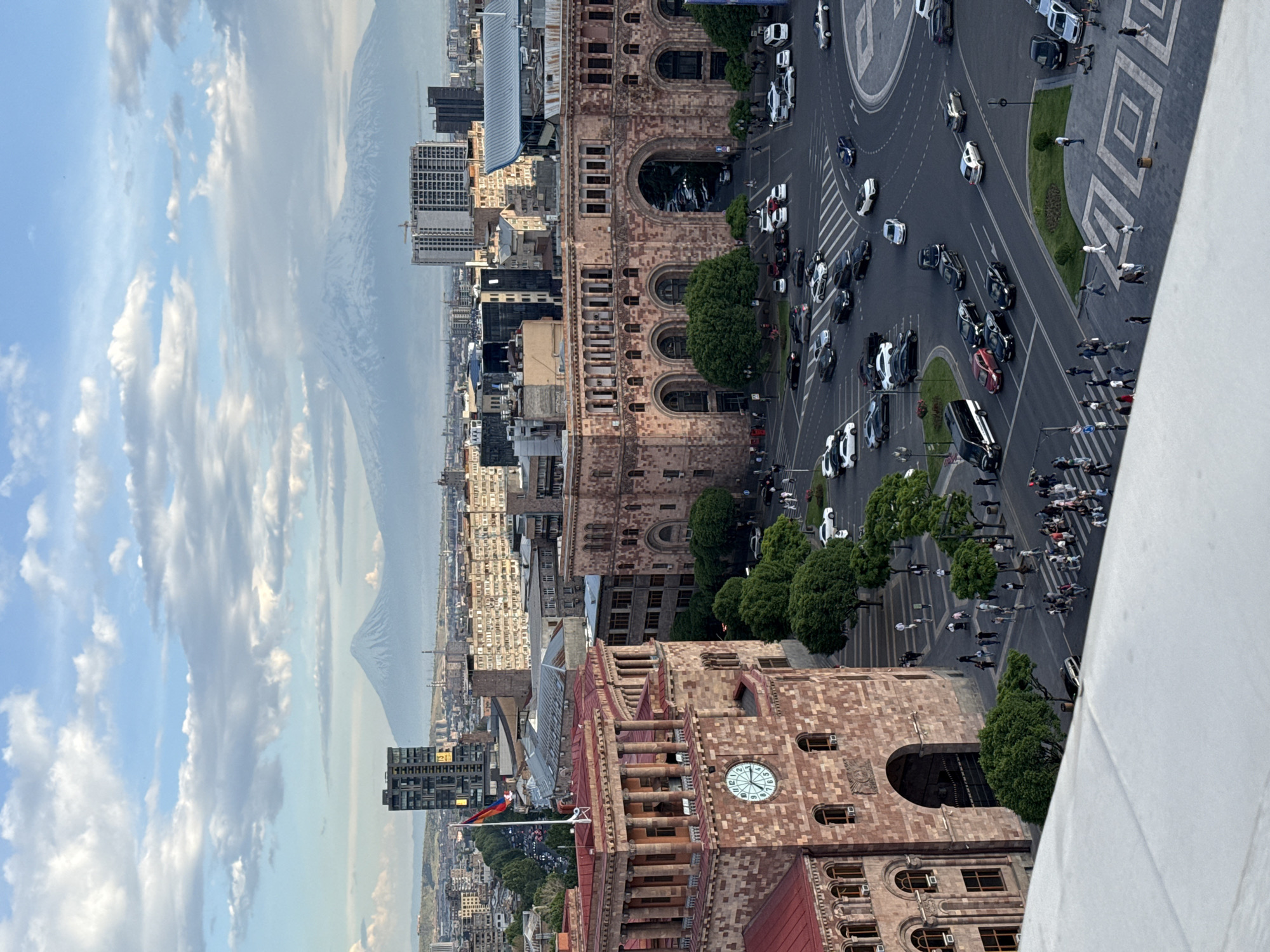 The image shows an aerial view of Yerevan, Armenia, focusing on Republic Square. The square has heavy traffic on the roundabout and sidewalks lined with trees. Pedestrians are visible in large numbers, some crossing the street. Buildings in the background, including the National Gallery, are visible, with Armenian flags on the clock tower of the government building. In the distance, Mount Ararat appears on the horizon. The sky is partly cloudy.