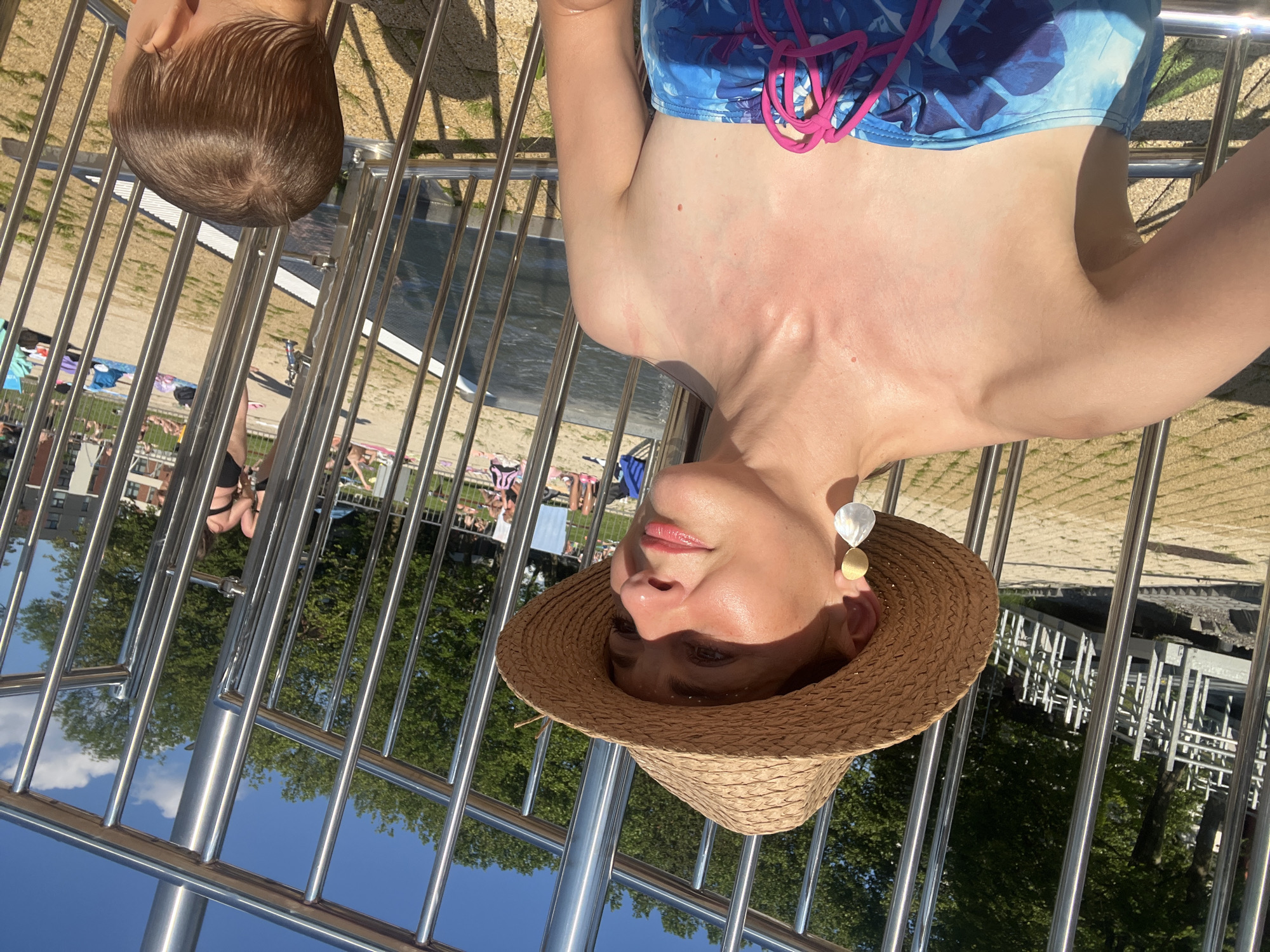 In an outdoor setting with a clear blue sky, a woman wearing a straw hat and a blue and white floral swimsuit is taking a selfie. She has blonde hair and is wearing large, teardrop-shaped earrings. The woman is seated outdoors, with a metal railing in the foreground. In the background, several people are visible sunbathing and relaxing by a pool area. There is also greenery from trees. Partially visible in the lower right corner of the image is the back of a young child's head, with wet brown hair. The scene appears to be a sunny day at a public pool or beach area.